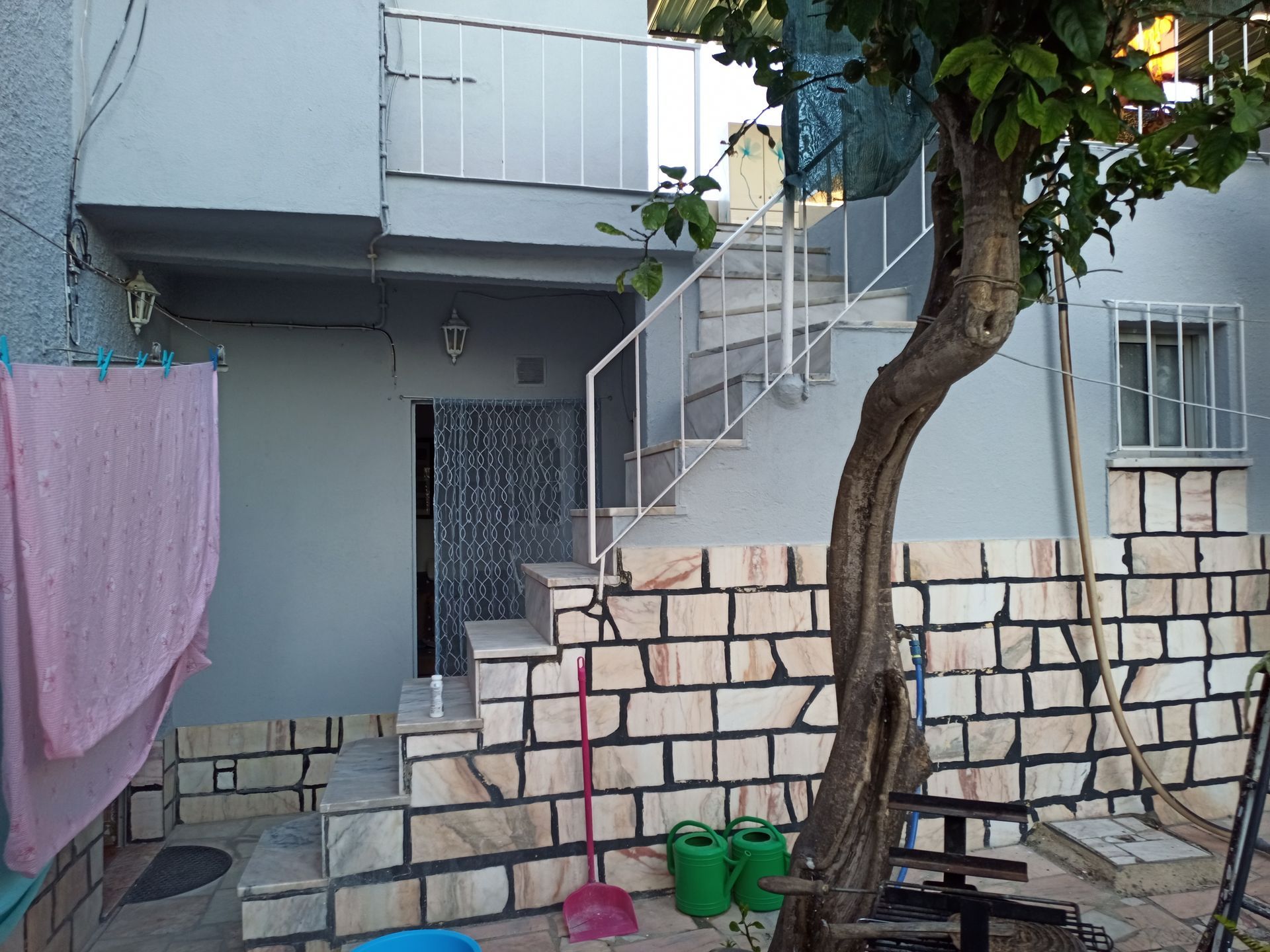 Exterior de um prédio com escadaria, árvore e roupa estendida. Paredes cinza-claro, fachada de pedra e corrimão de metal.