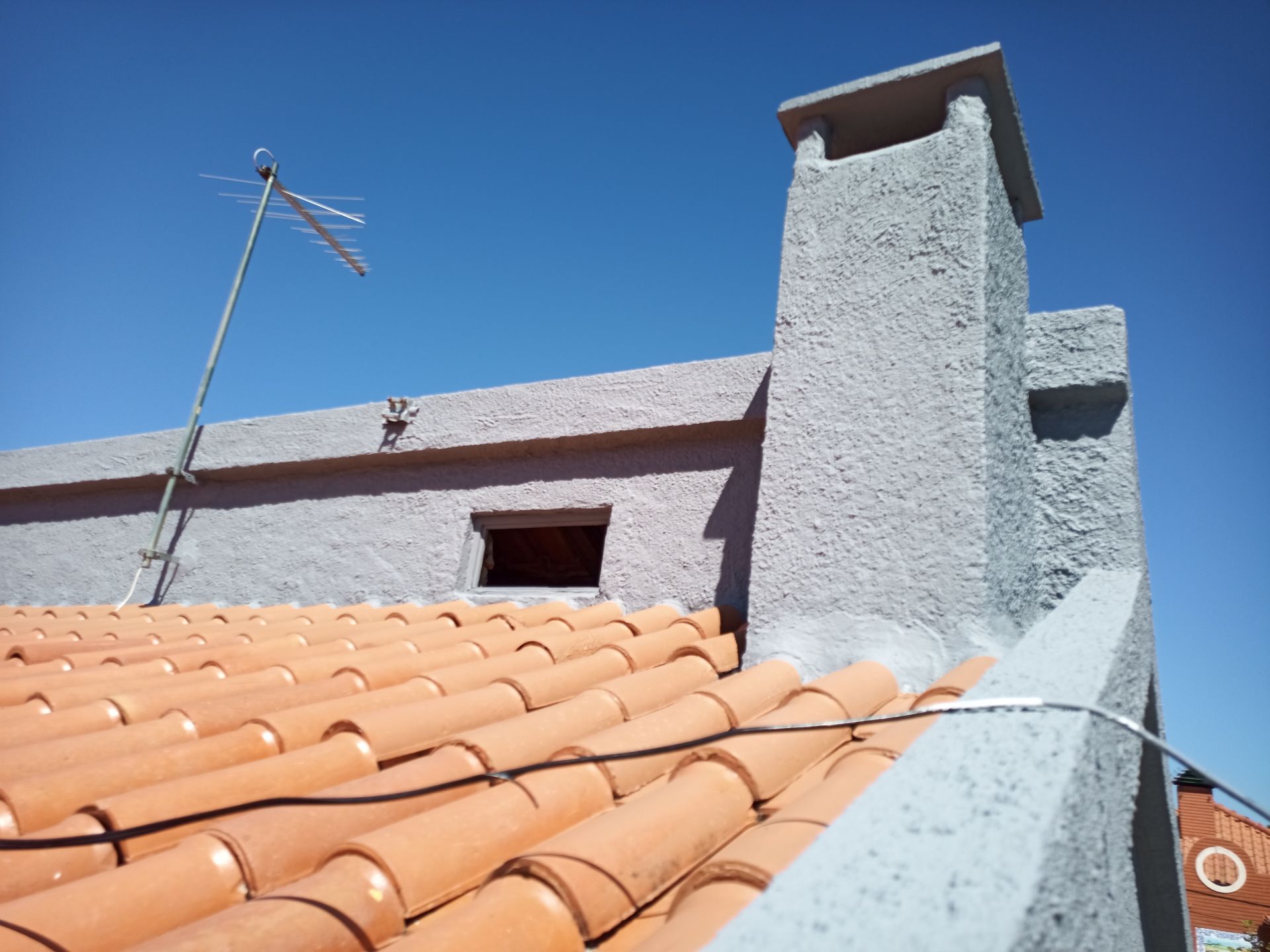 Telhado de telhas vermelhas com antena, chaminé e edifício de estuque cinza contra um céu azul claro.