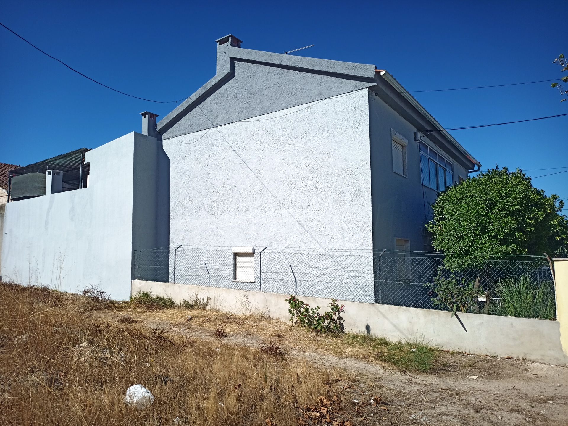 Casa pintada de cinza com uma pequena janela, chaminé e uma pequena cerca, tendo como pano de fundo um céu azul e grama seca e alta.