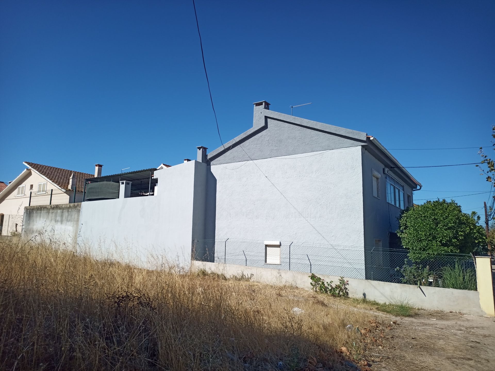 Casa de estuque cinza com chaminé e pequena janela, vista de uma encosta gramada contra um céu azul.