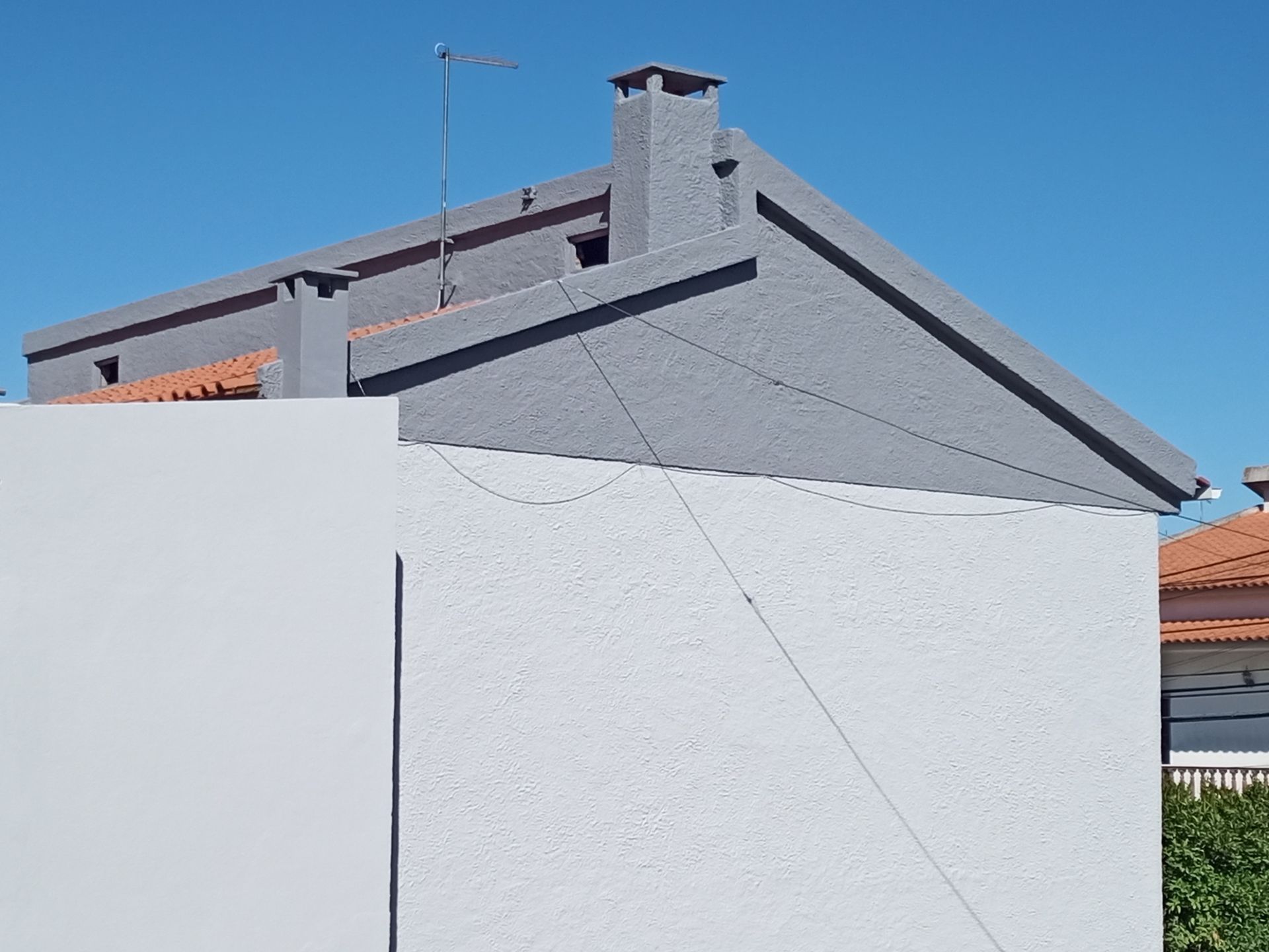 Prédio com telhado cinza e fachada branca contra um céu azul claro. Uma chaminé e uma antena são visíveis.
