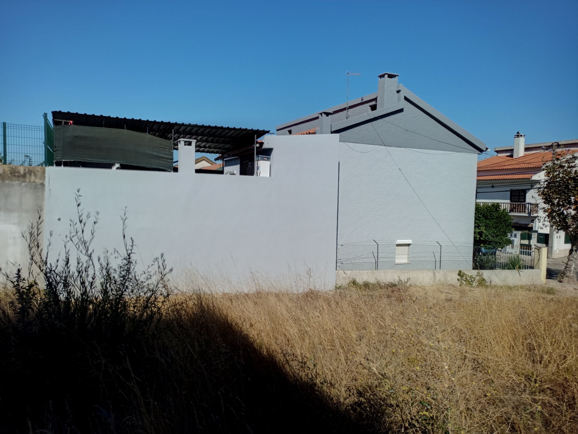 Prédio pintado de cinza com toldo escuro em um dia ensolarado. Grama alta e seca em primeiro plano.