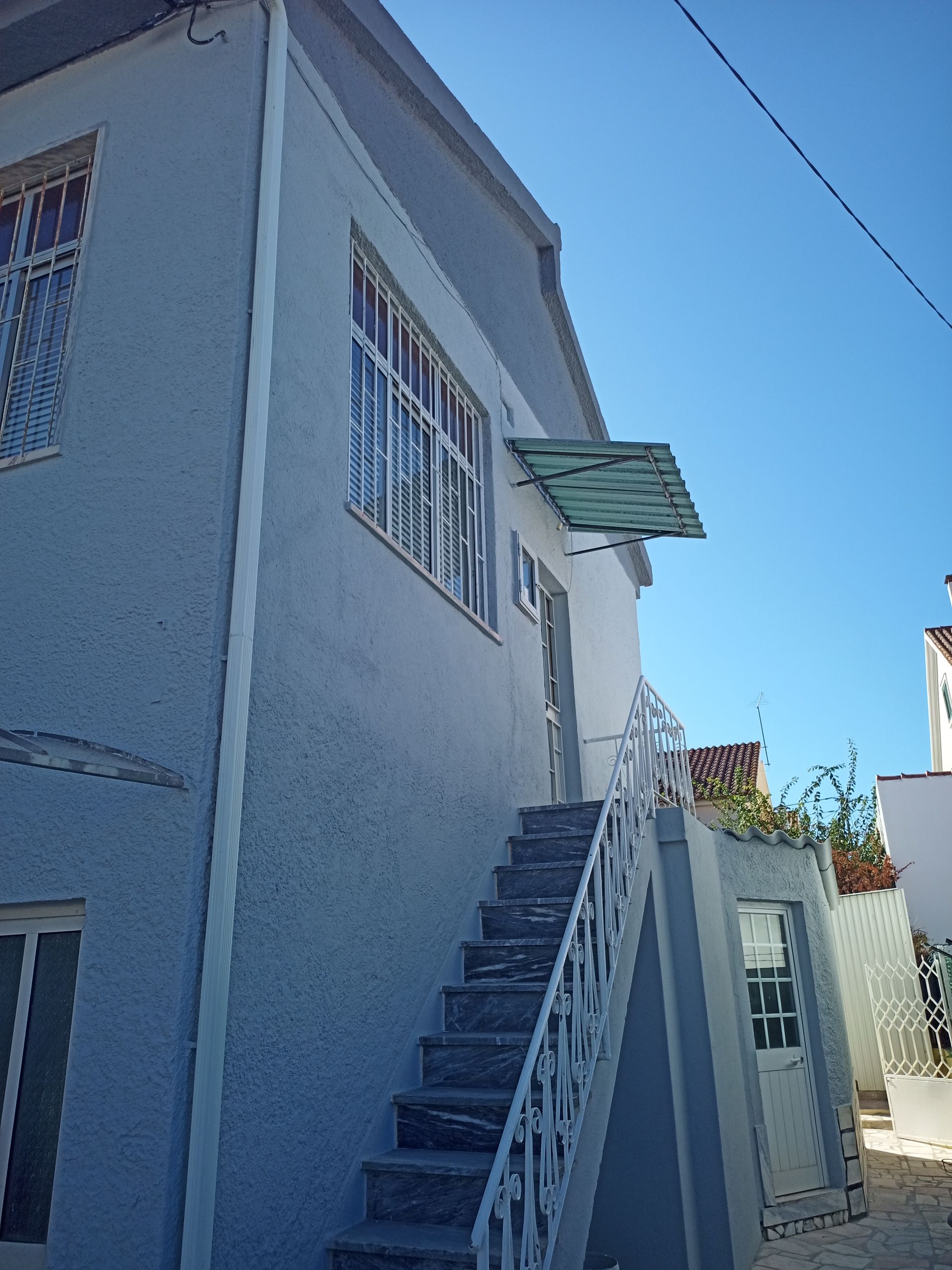 Prédio cinza de dois andares com escada externa que leva a uma porta; grades de metal nas janelas; céu azul.