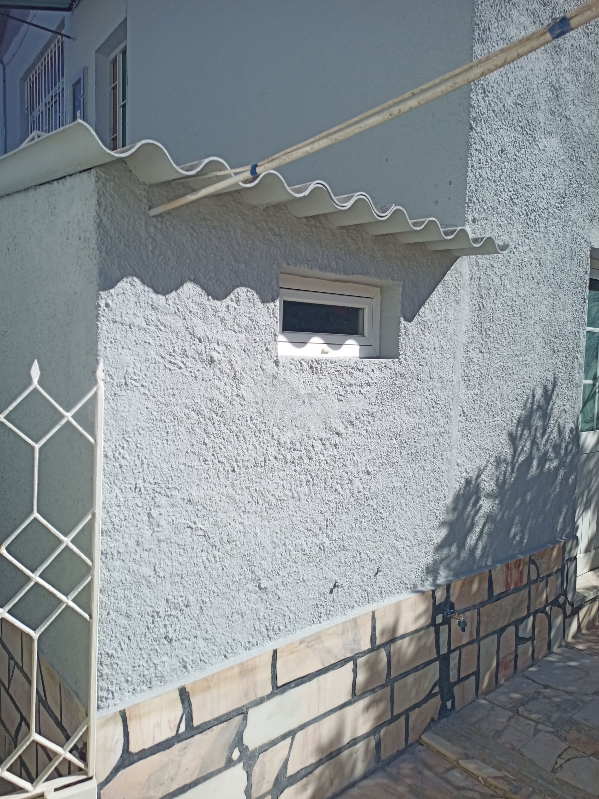 Vista exterior de um edifício com uma parede cinzenta texturizada, uma base de tijolos e um toldo ondulado sobre uma pequena janela.