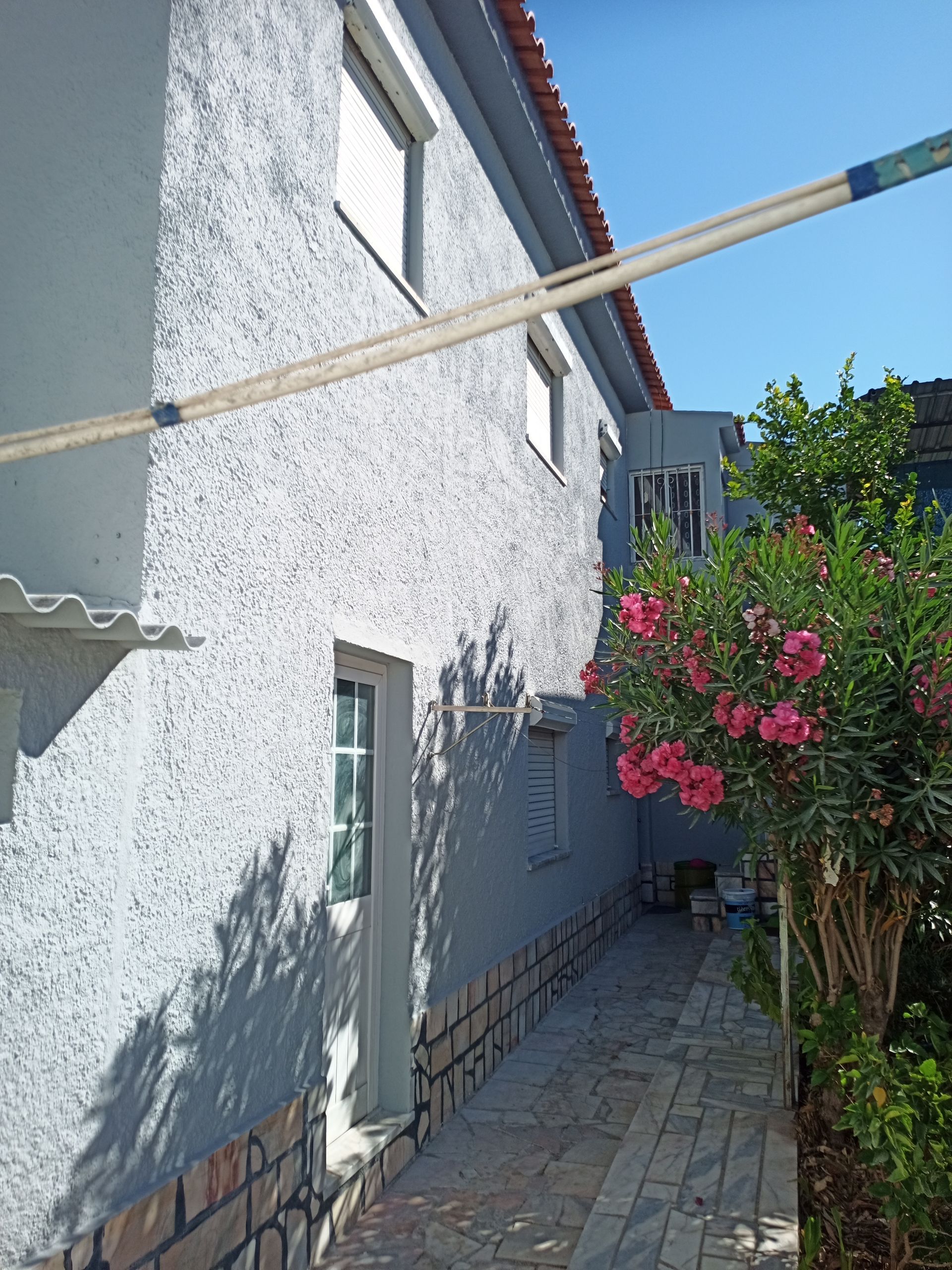 Casa de estuque cinza com caminho de tijolos e arbusto florido rosa sob um céu azul claro.