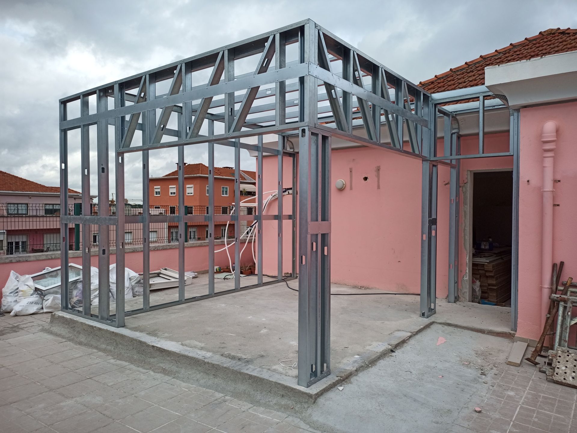 Estrutura metálica de um anexo em construção em um telhado, com uma parede rosa ao fundo.