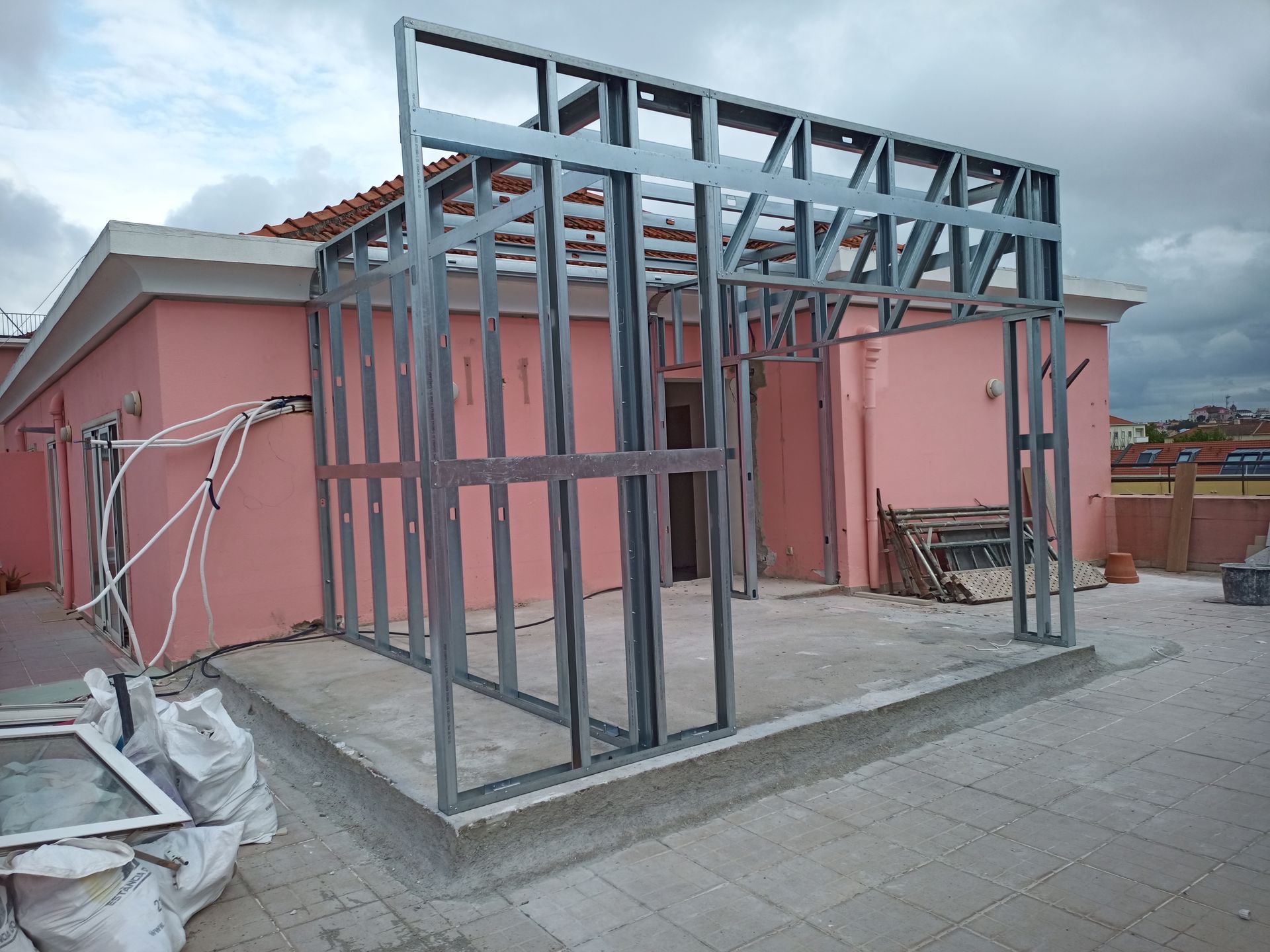 A estrutura metálica externa de um edifício está sendo construída sobre uma laje de concreto, perto de um prédio rosa.