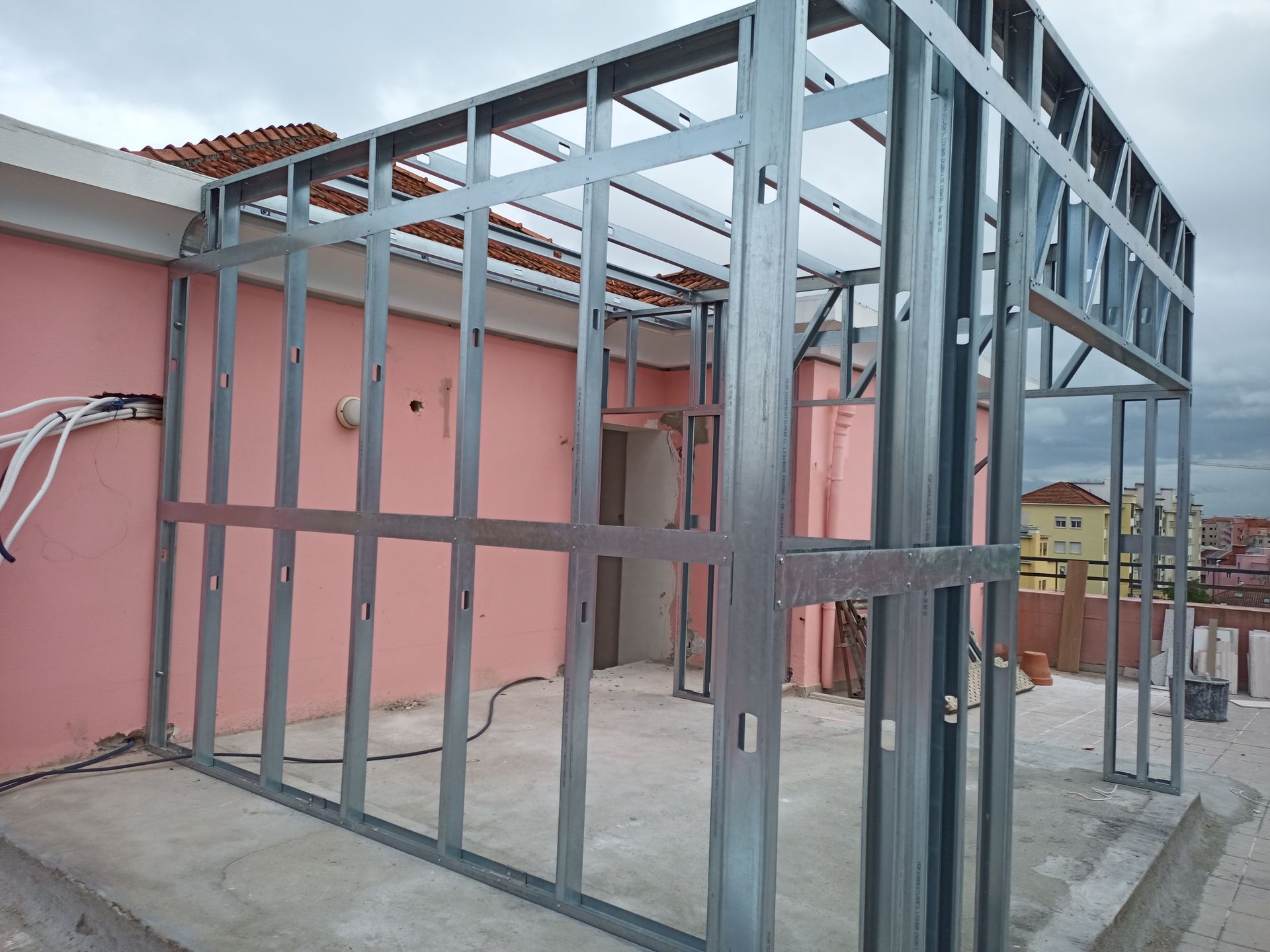 A estrutura metálica das paredes externas de um edifício está sendo construída em um telhado com paredes rosas e céu nublado.