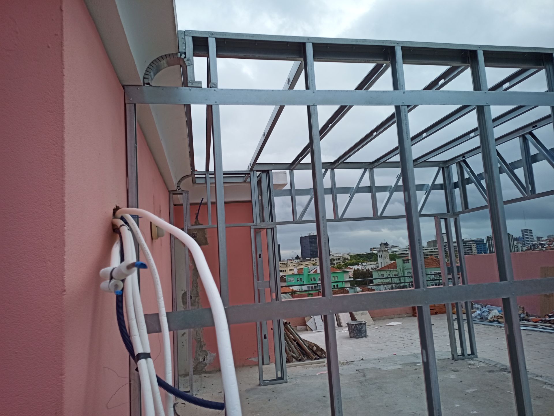 Estrutura metálica em um telhado, com parede rosa, encanamento e paisagem urbana ao fundo.