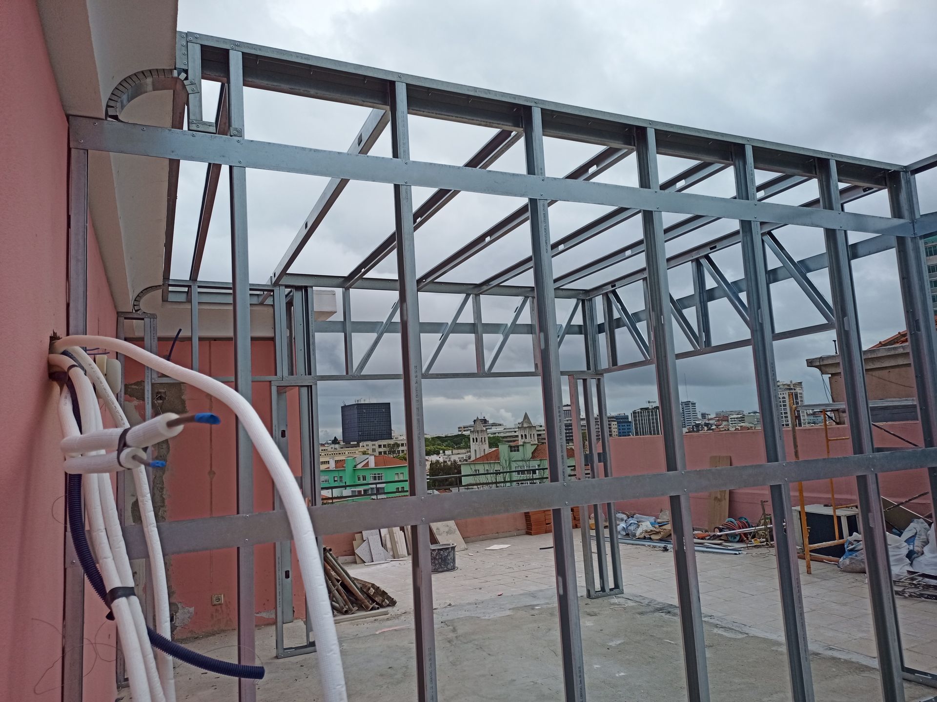 Estrutura metálica no telhado com parede rosa, tubulações azuis e brancas e vista para a cidade.