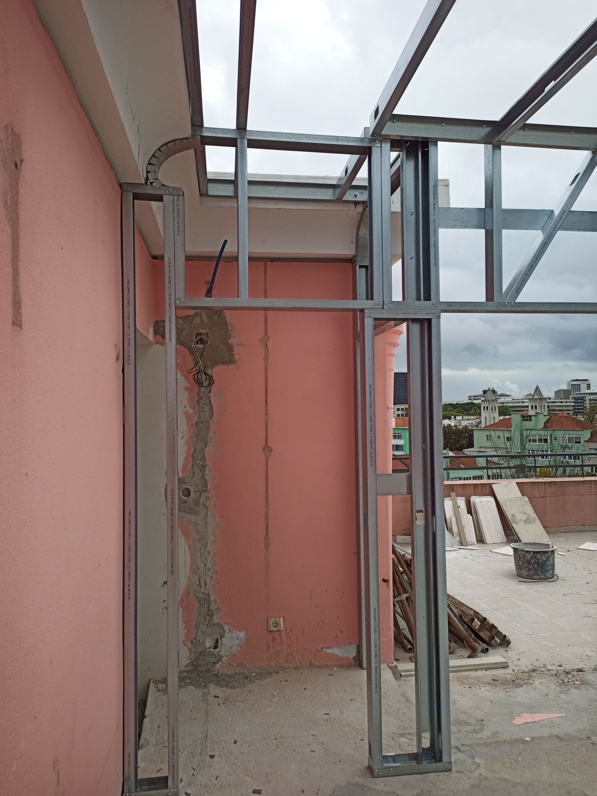 Estrutura metálica de uma construção em andamento; parede rosa e ambiente urbano ao fundo.