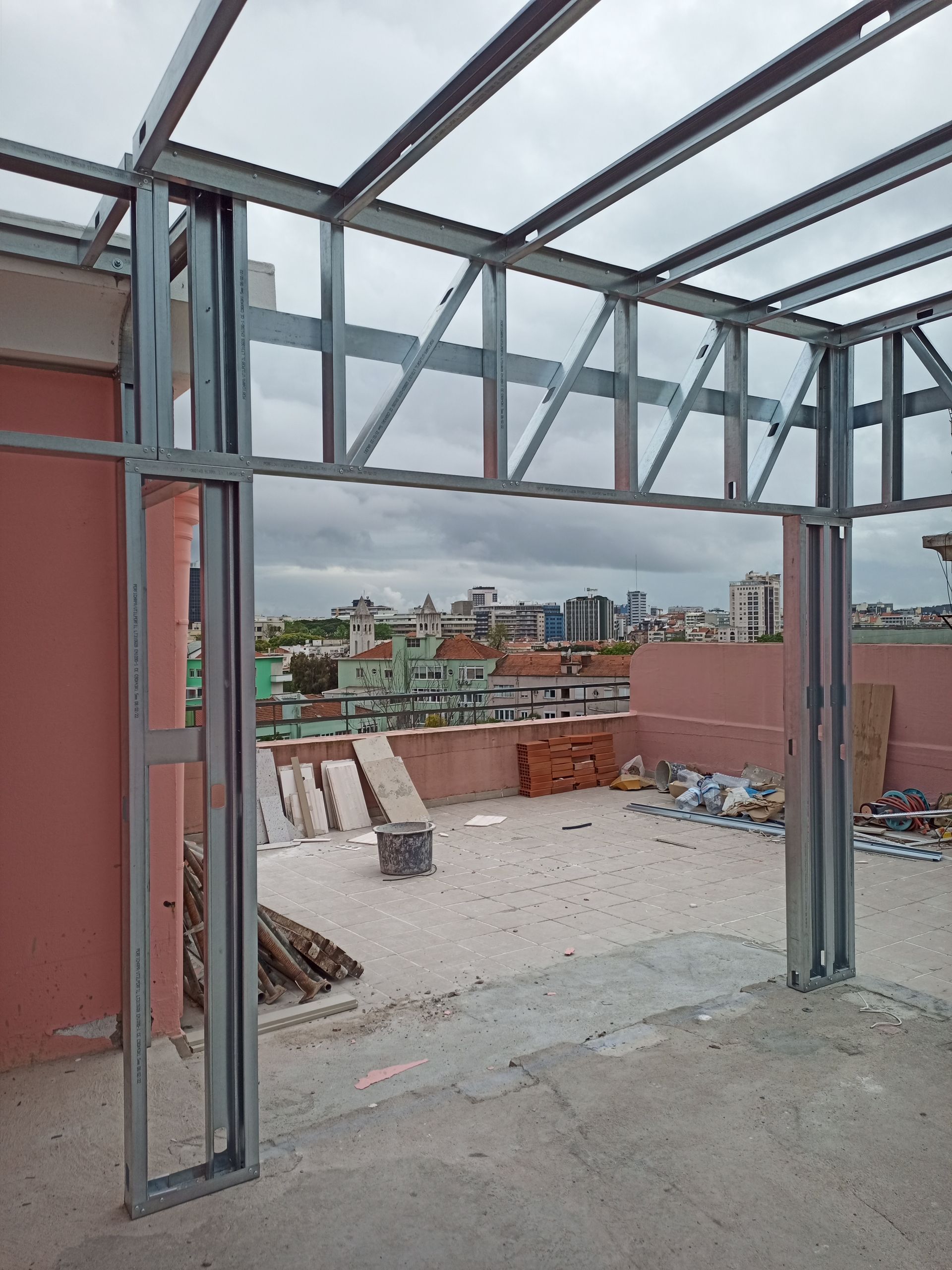 Estrutura metálica para o telhado e a entrada de um edifício, com vista para a paisagem urbana sob um céu nublado.