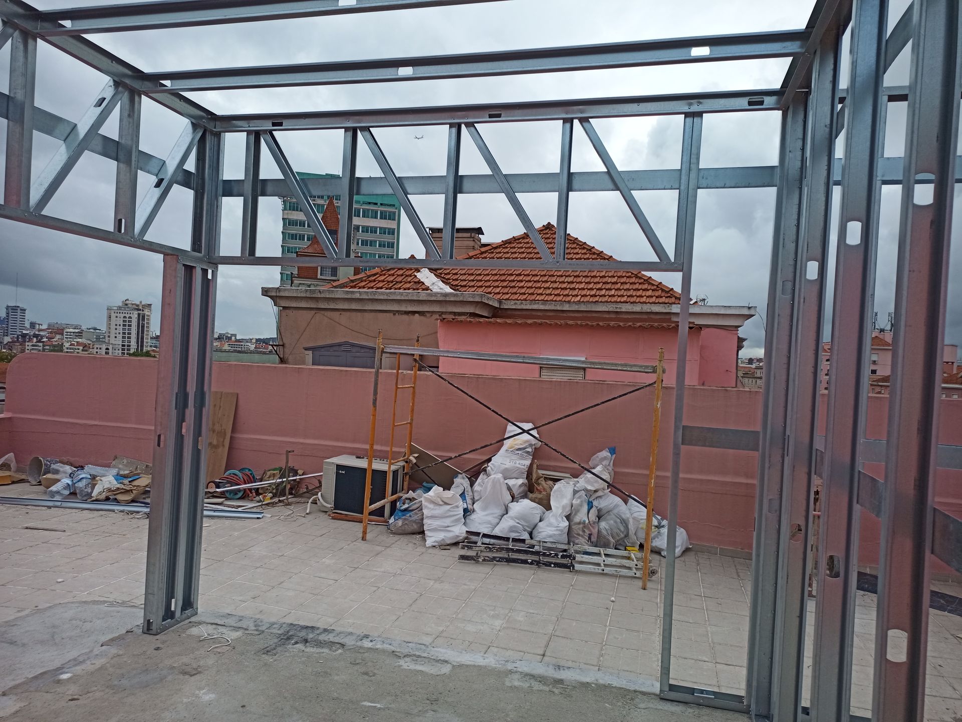 Estrutura de aço em um telhado com parede rosa, sacos de materiais e cidade ao fundo.