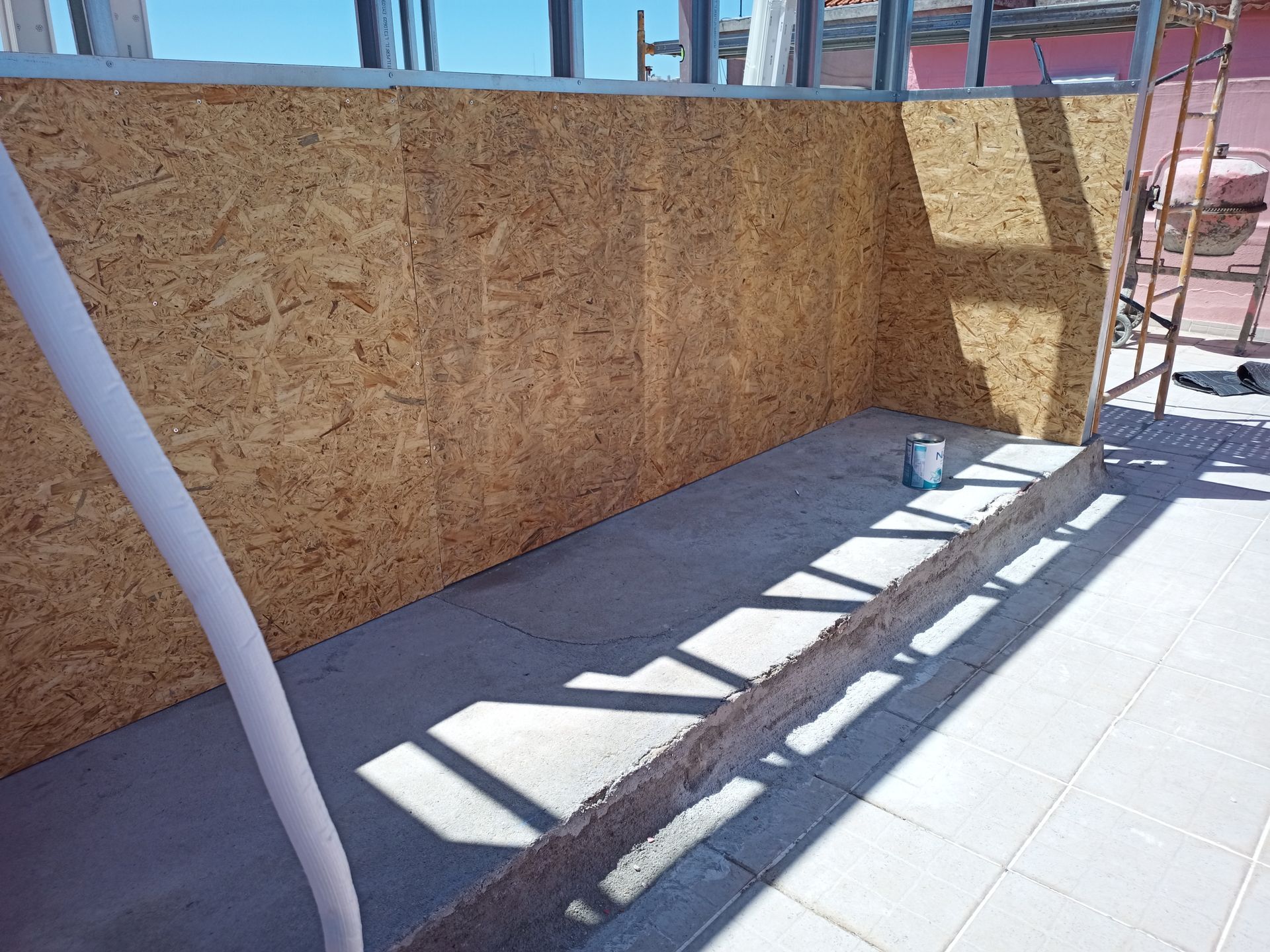 Canteiro de obras com painéis de madeira fixados em uma estrutura metálica. Uma mangueira de cor clara percorre uma base de concreto.