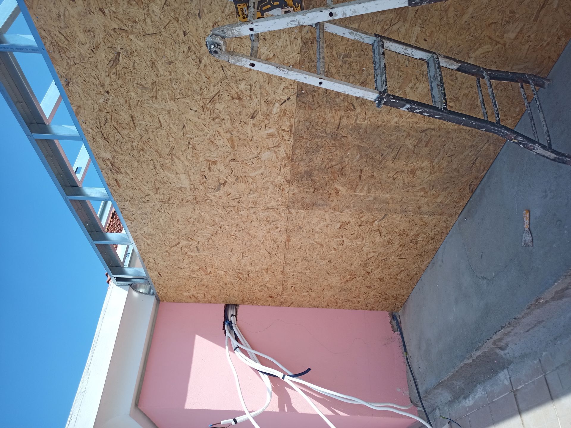 Teto em construção: placas OSB, estrutura metálica, escada, isolamento rosa, fios, parede de concreto, céu azul.