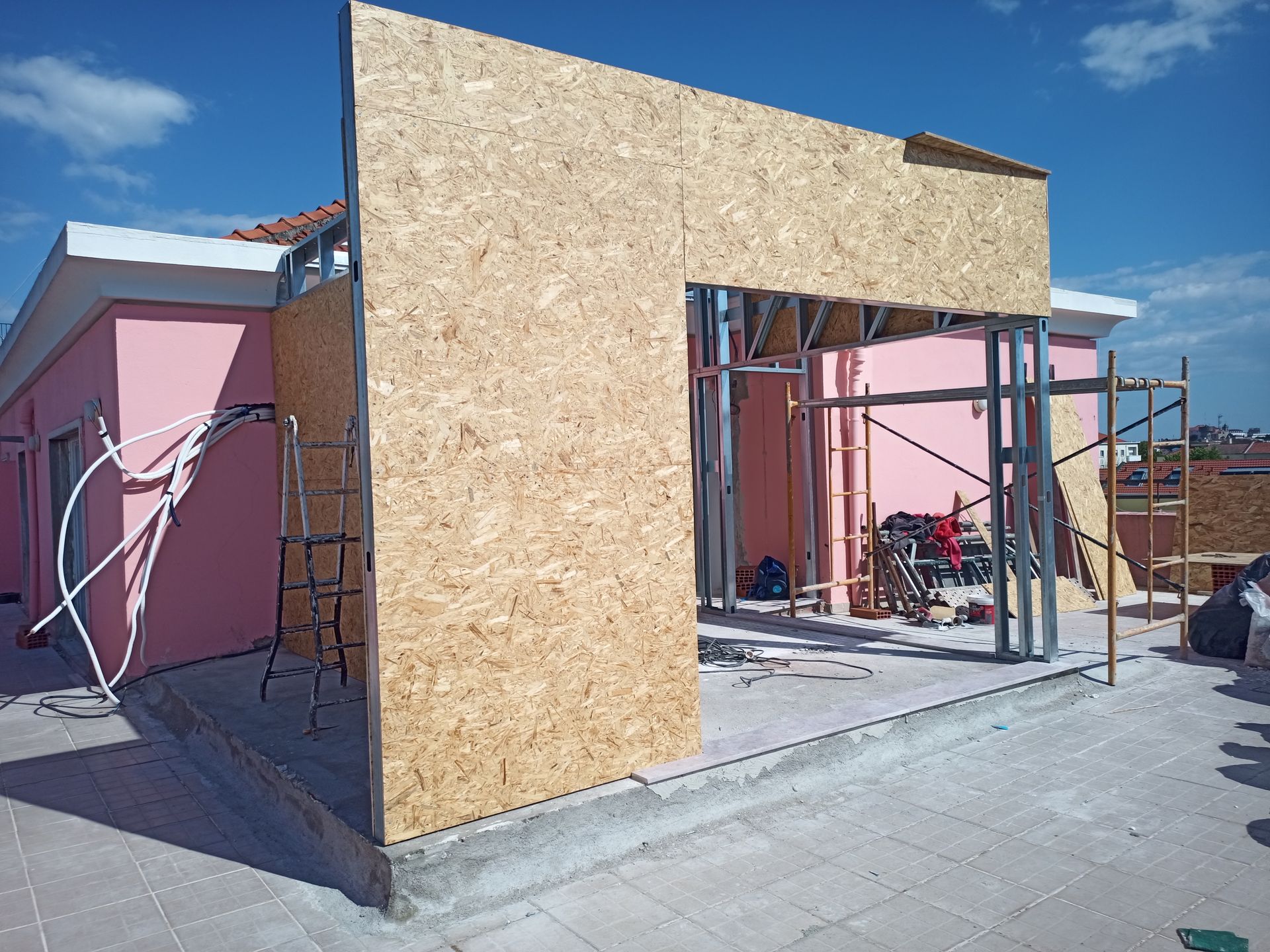 Construção da parede externa de um edifício com painéis de madeira orientada (OSB) e estrutura de aço em um telhado, com um prédio rosa ao fundo.