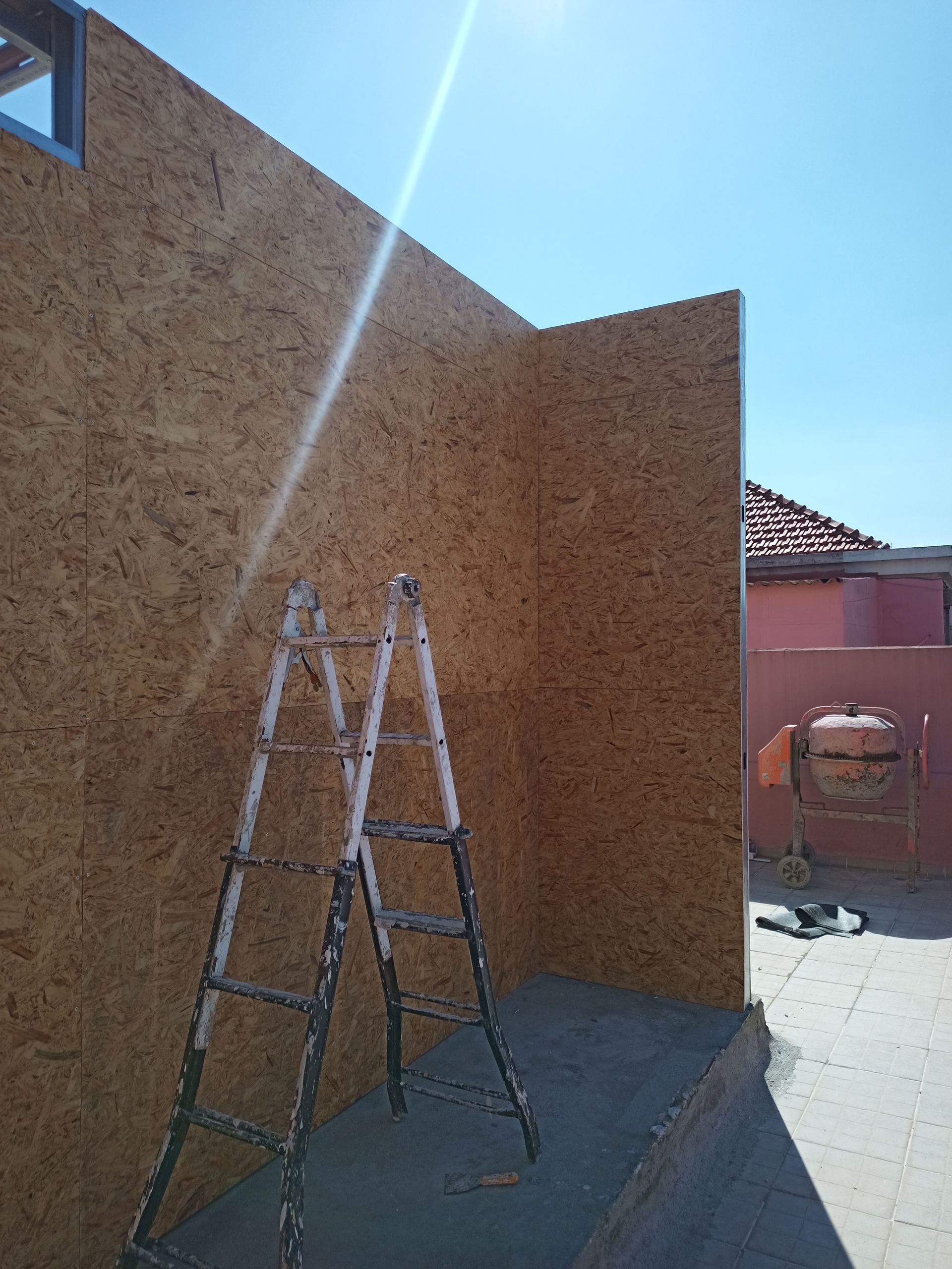 Escada encostada em uma parede de madeira em construção, com céu azul e vista parcial de um edifício.