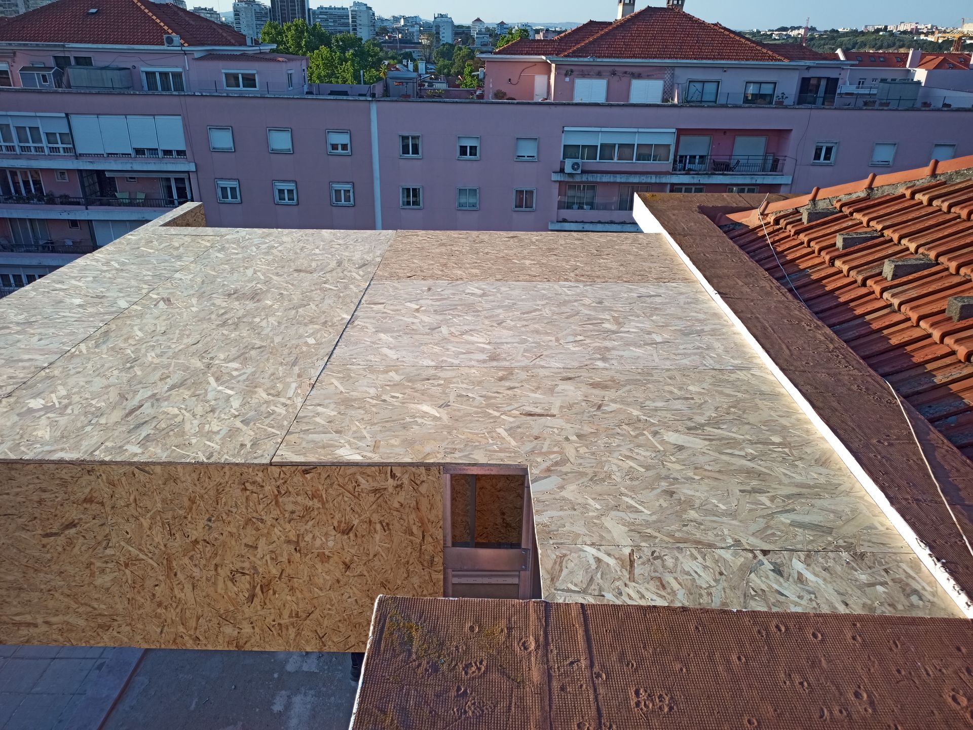 Telhado revestido com OSB, com estrutura parcialmente acabada, telha vermelha e prédios da cidade ao fundo.