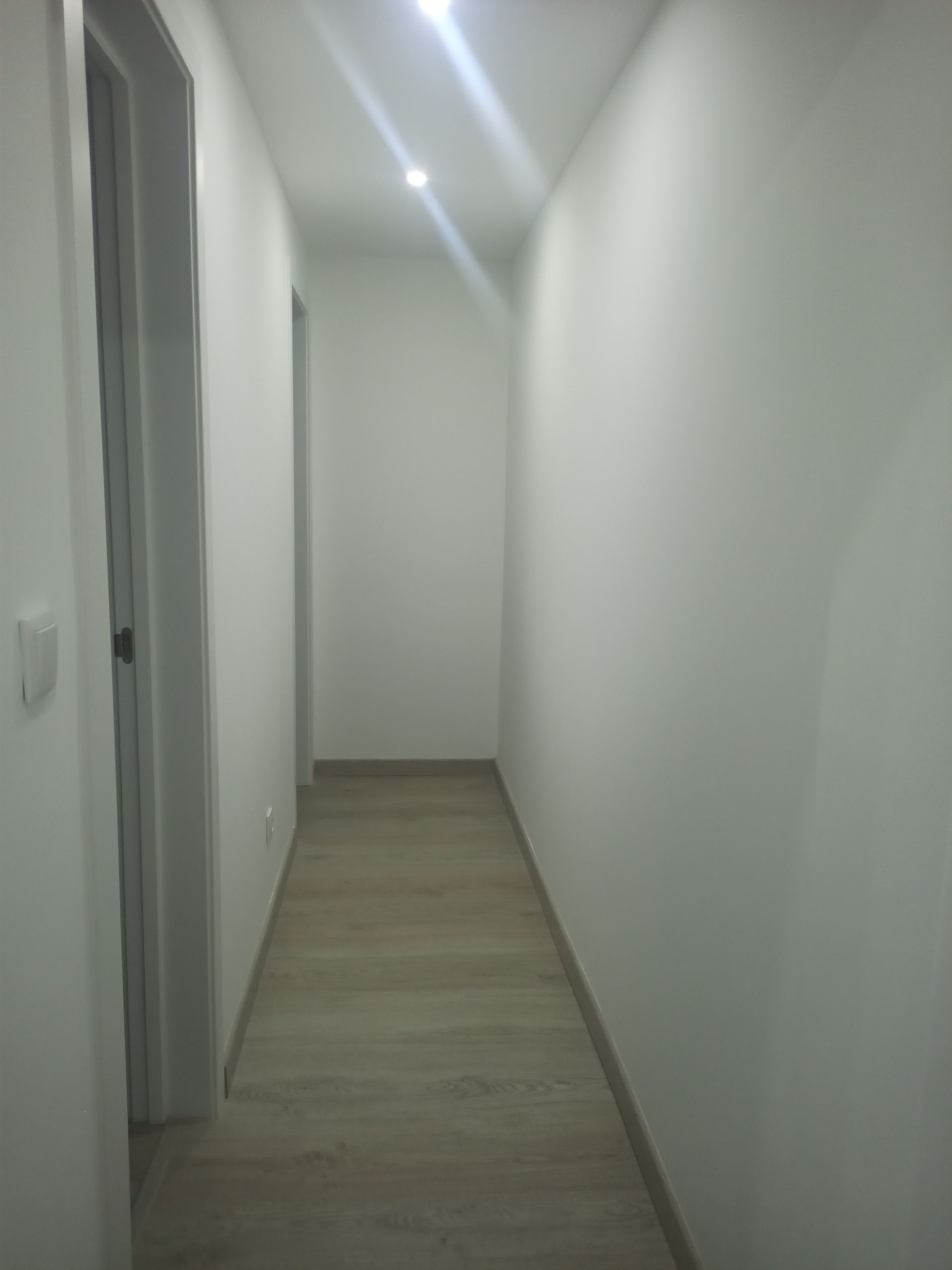 Corredor estreito com paredes brancas, piso de madeira clara e iluminação no teto.