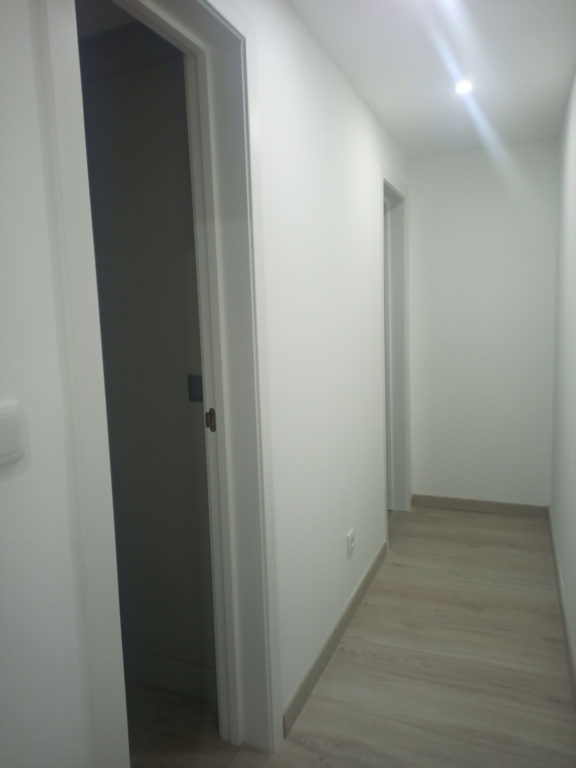 Corredor estreito com paredes brancas e piso de madeira, duas portas à esquerda e uma em frente.