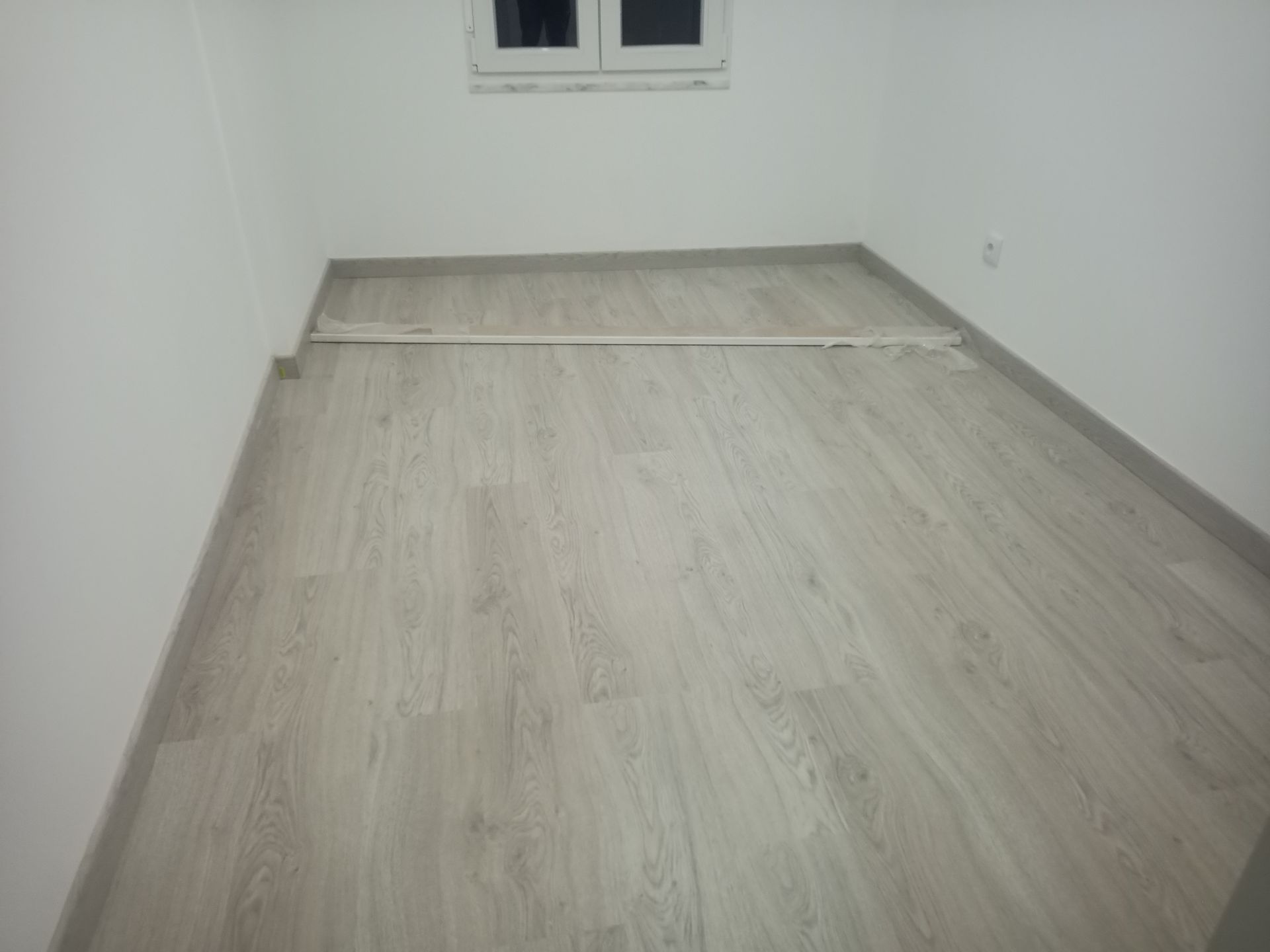 Quarto vazio com piso laminado imitando madeira clara, paredes brancas e uma pequena janela.