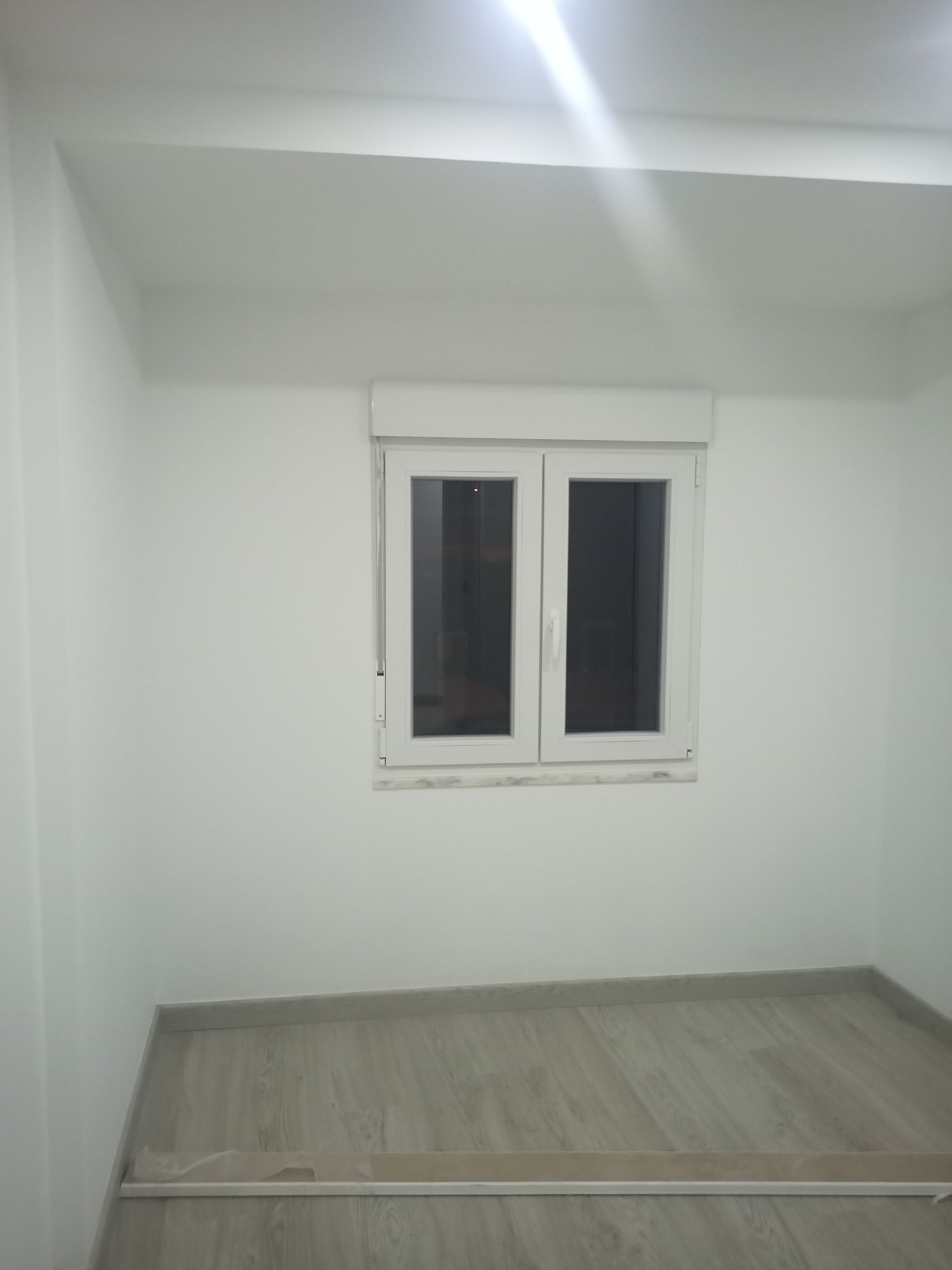 Quarto branco vazio com janela, piso que imita madeira clara e uma faixa de madeira no chão.