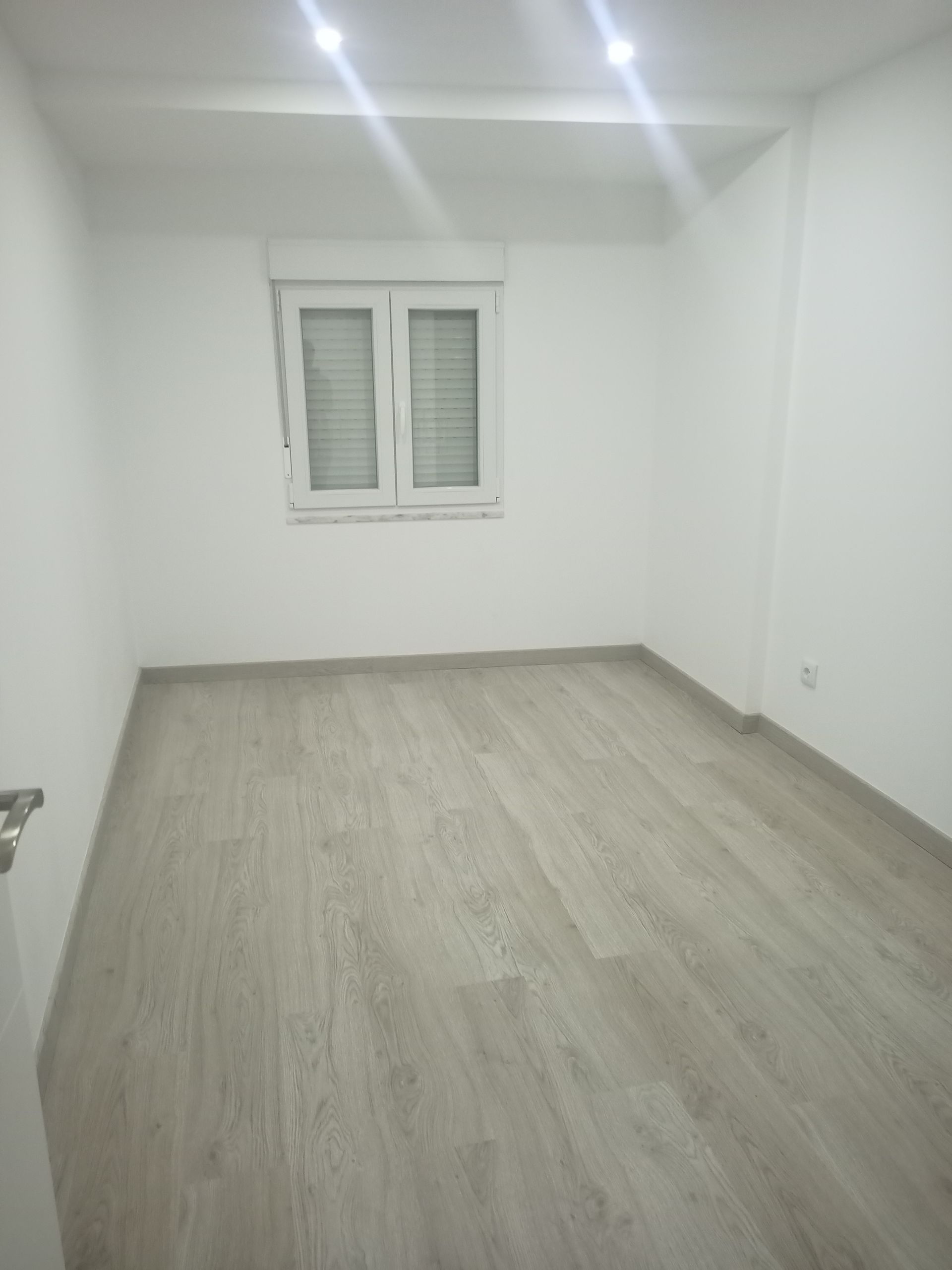 Quarto branco vazio com piso que imita madeira clara, uma janela e luzes no teto.