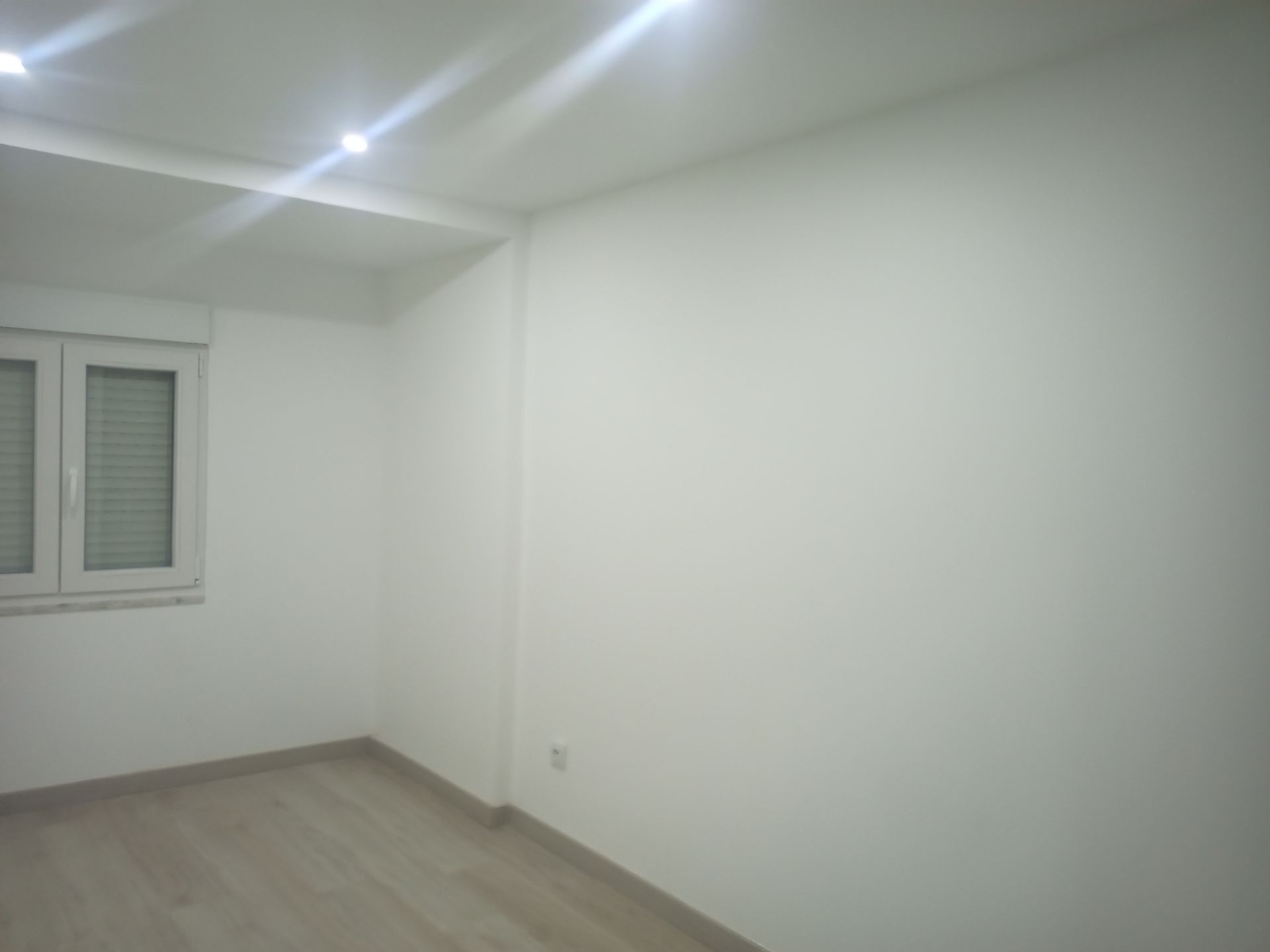 Quarto vazio com paredes brancas, janela com persianas e piso de madeira clara.