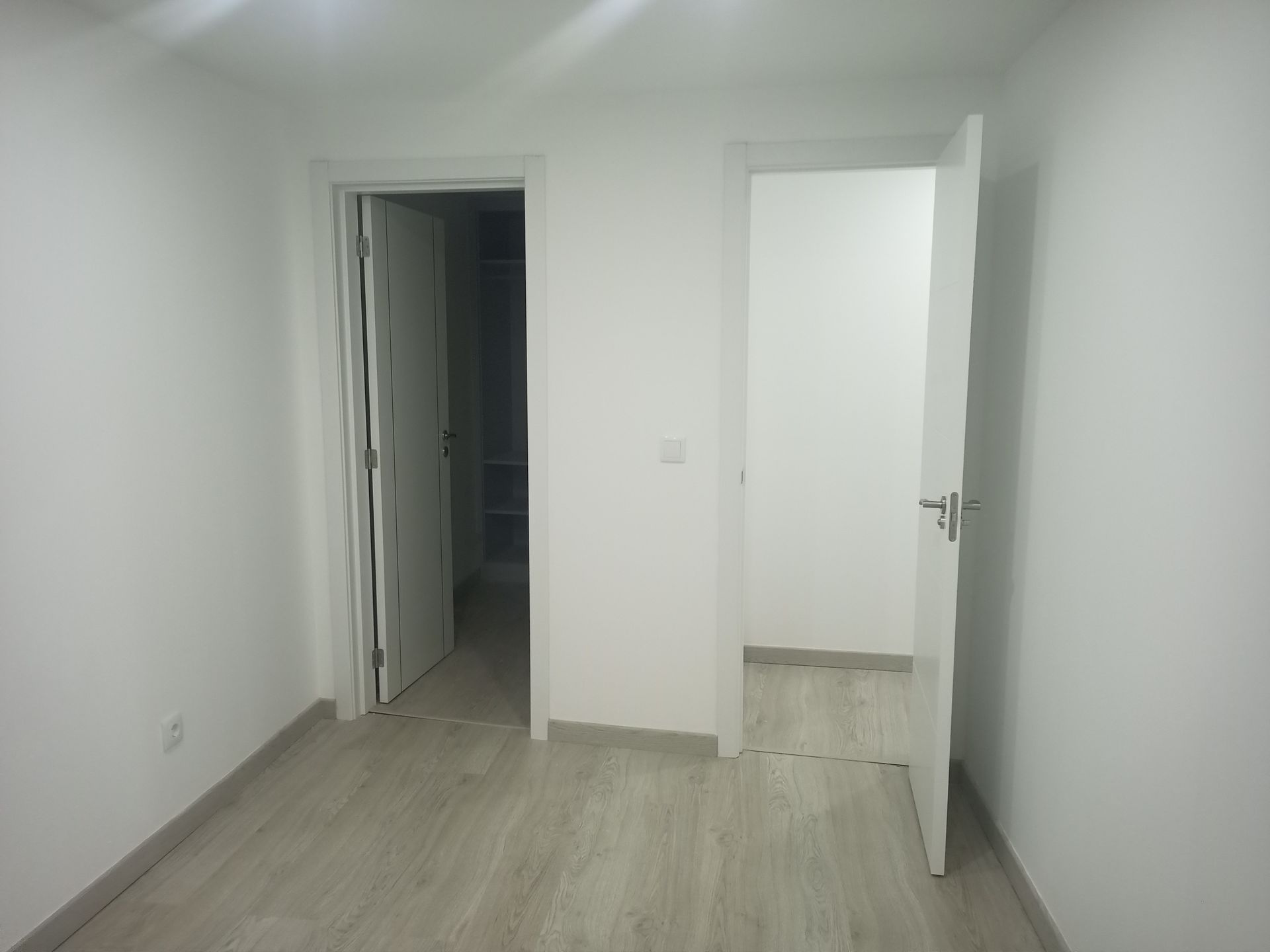 Quarto vazio com duas portas abertas, paredes brancas e piso de madeira clara.