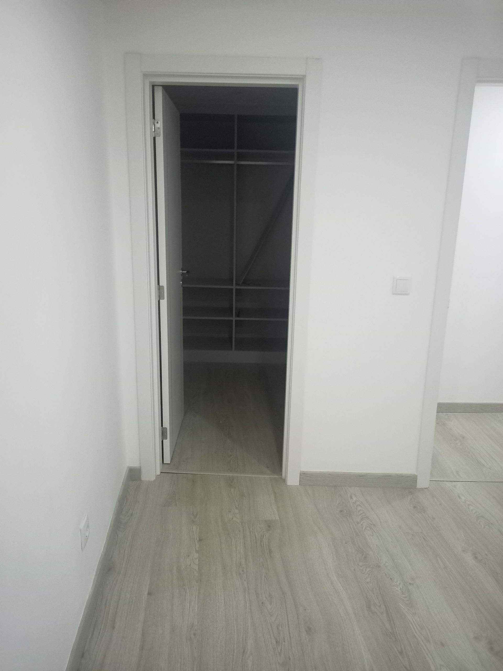 Uma porta dá acesso a um closet com prateleiras e um cabideiro. Paredes brancas, piso cinza.