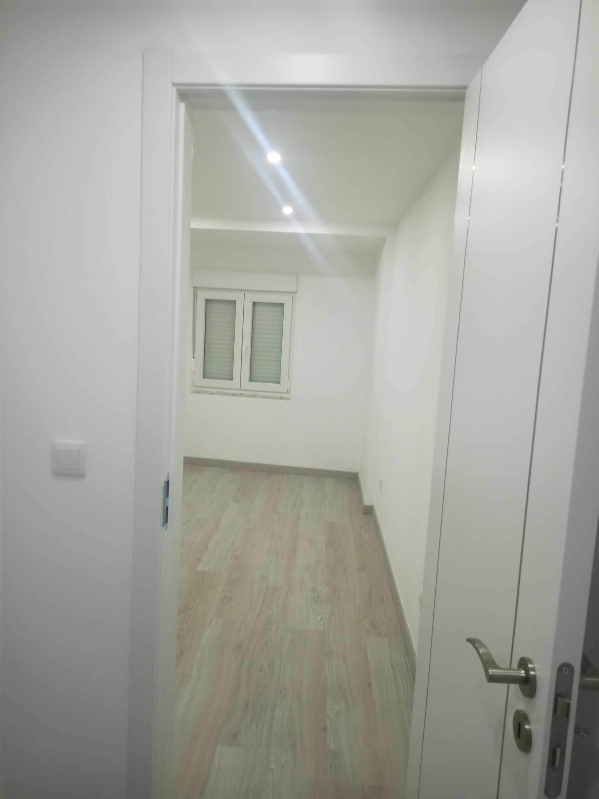 Vista de um quarto branco com piso que imita madeira através de uma porta. Há uma janela ao fundo.