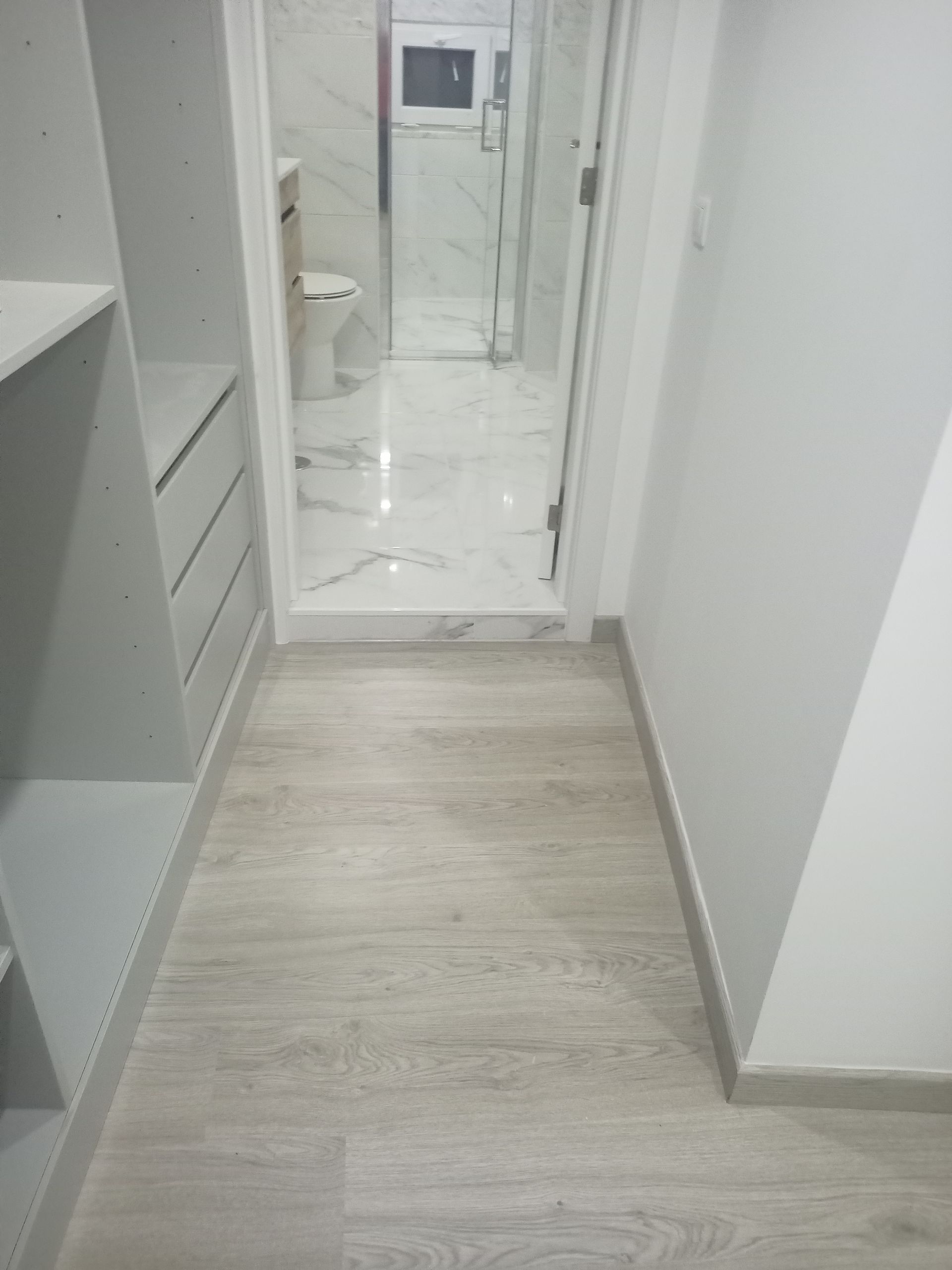 Corredor estreito com piso que imita madeira clara, que leva a um banheiro com azulejos brancos e box de vidro.