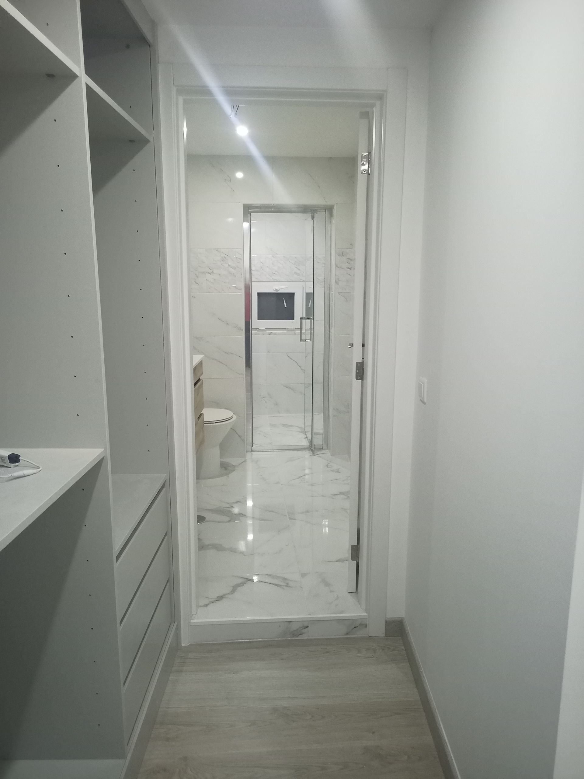 Corredor estreito com box de vidro no chuveiro, armário embutido, piso de mármore e paredes brancas.