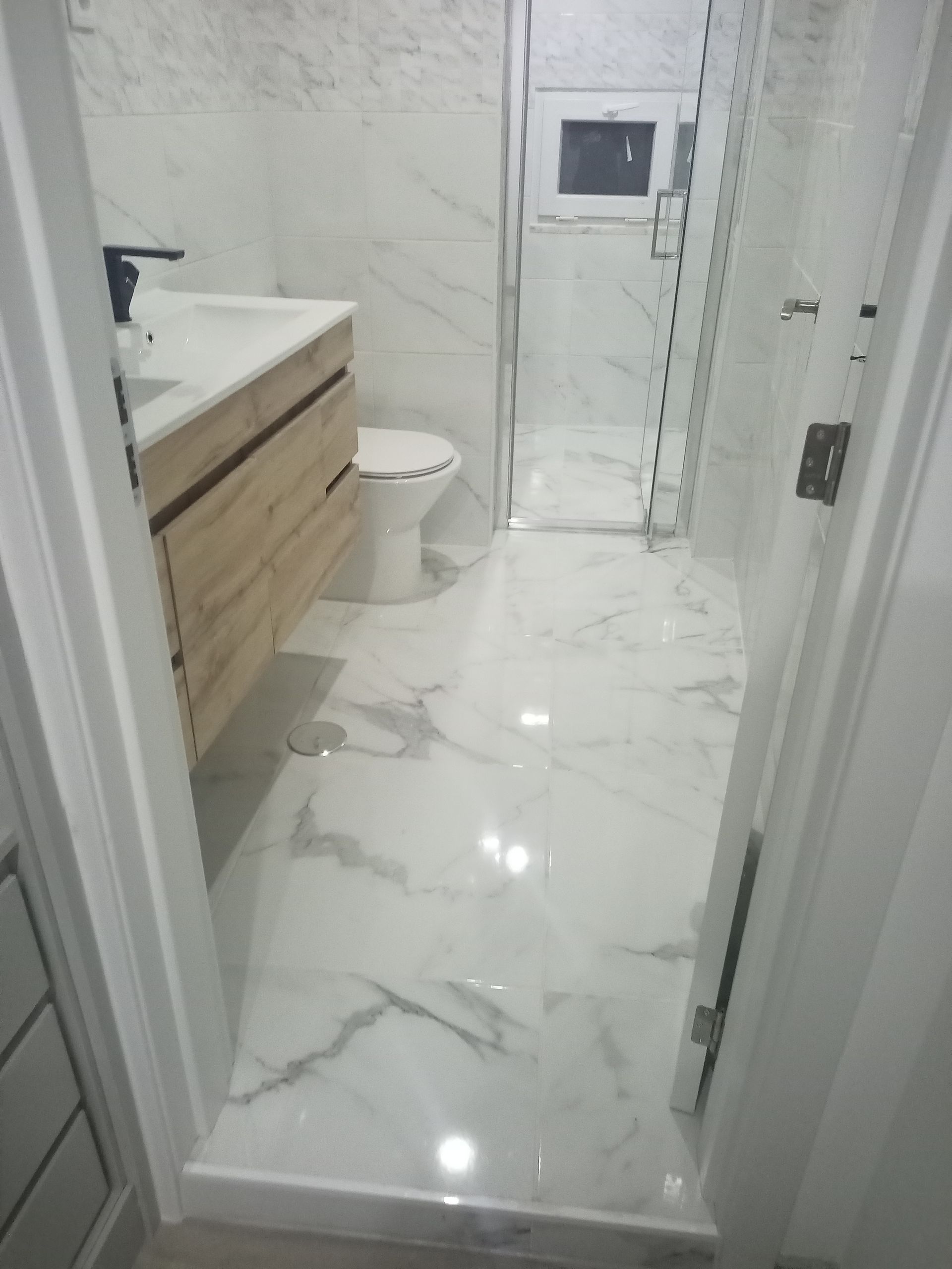 Banheiro com azulejos de mármore branco, pia de madeira, vaso sanitário e box de vidro.
