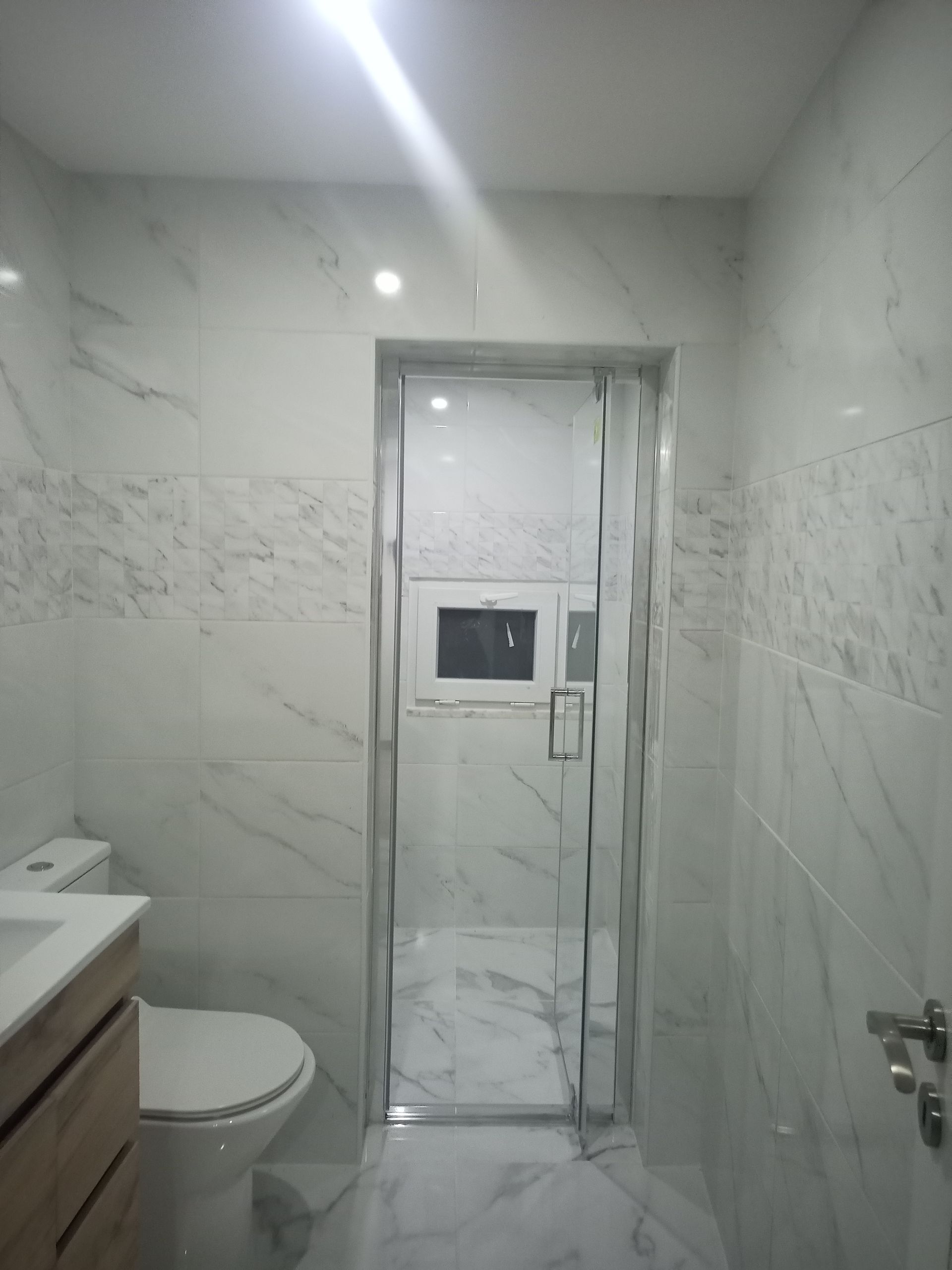 Banheiro com azulejos brancos, box de vidro, vaso sanitário e pia.