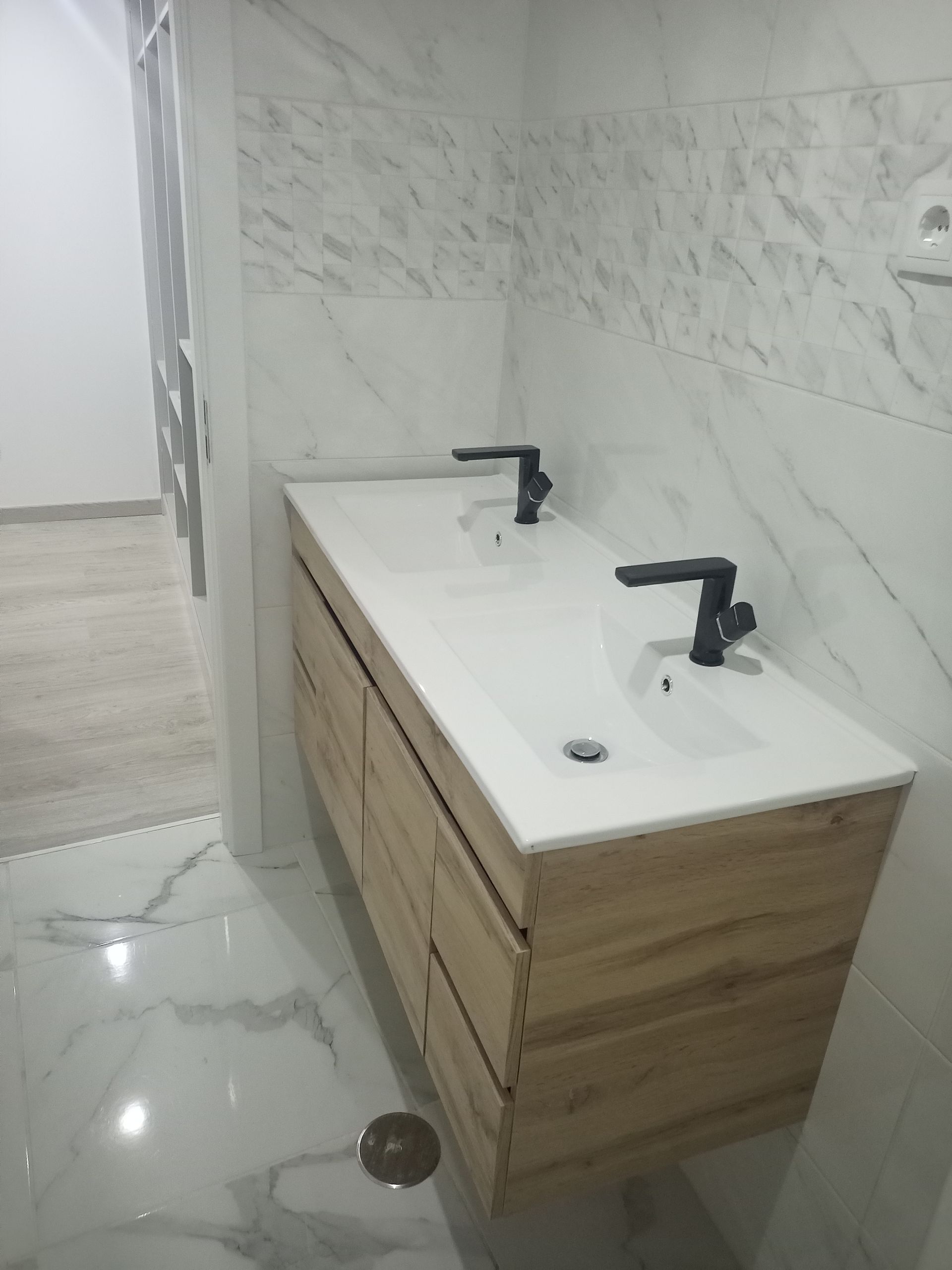 Banheiro com pia dupla; bancada branca e azulejo de porcelana, gavetas de madeira, torneiras pretas, paredes revestidas com azulejos imitando mármore.