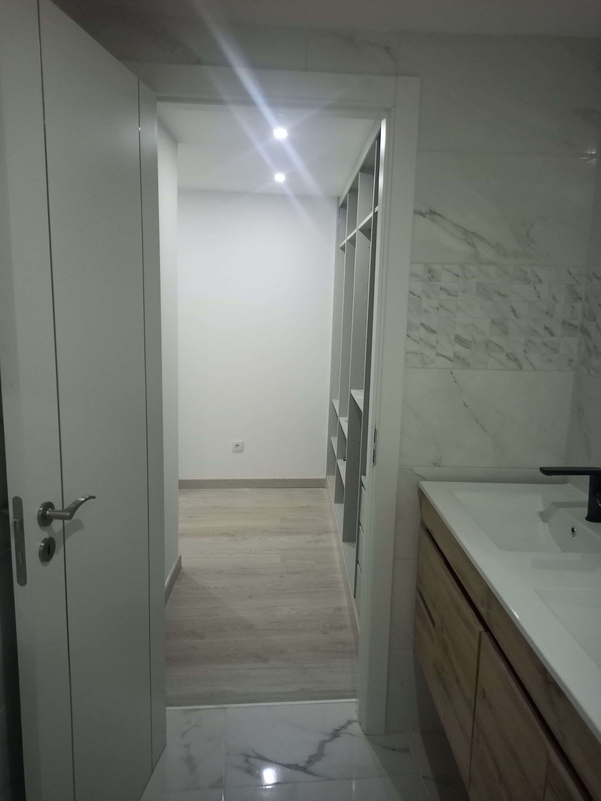 Interior de banheiro com porta aberta para um quarto. Paredes brancas, bancada de madeira e box de vidro estampado são visíveis.