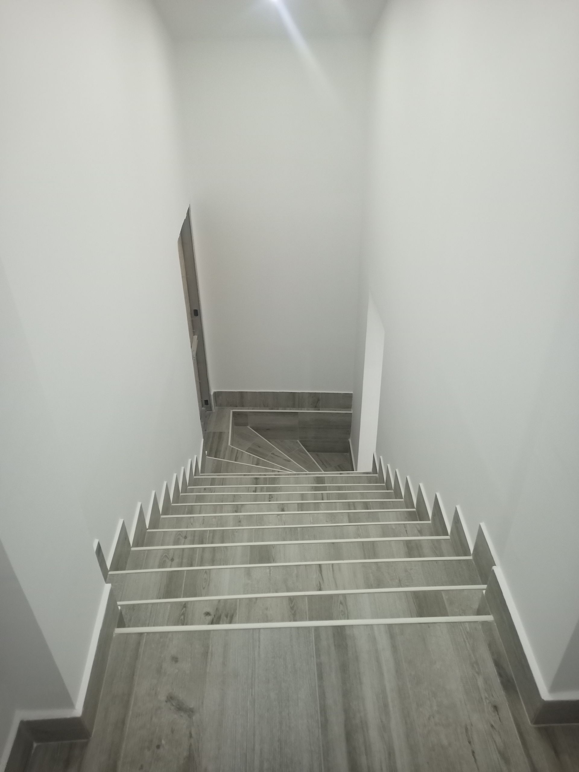 Escada com degraus de madeira cinza, paredes brancas e uma porta no topo.