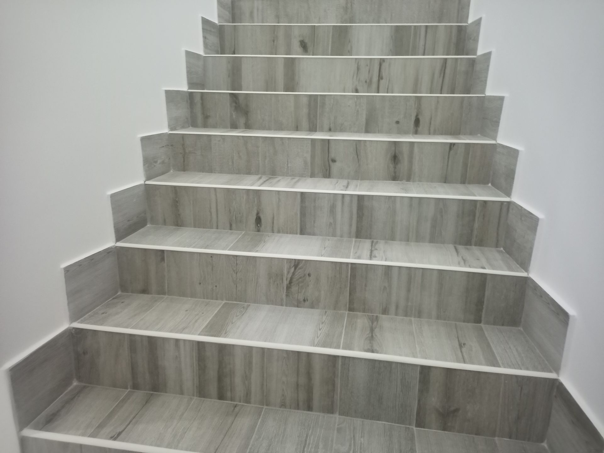 Escada com degraus revestidos com azulejos imitando madeira cinza e paredes brancas, subindo em direção ao andar superior.