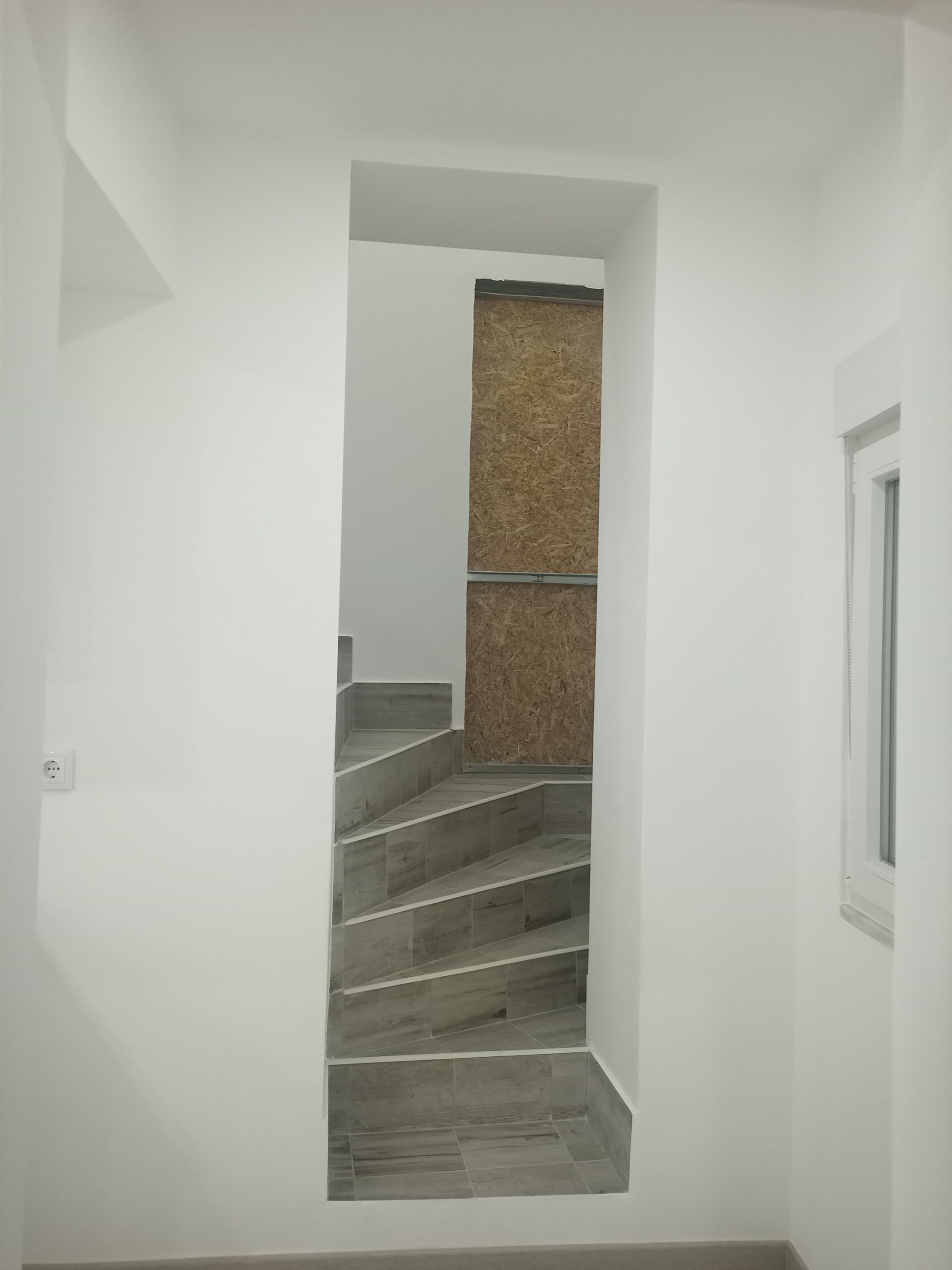 Escada vista através de uma abertura em uma parede branca; os degraus levam a uma porta.