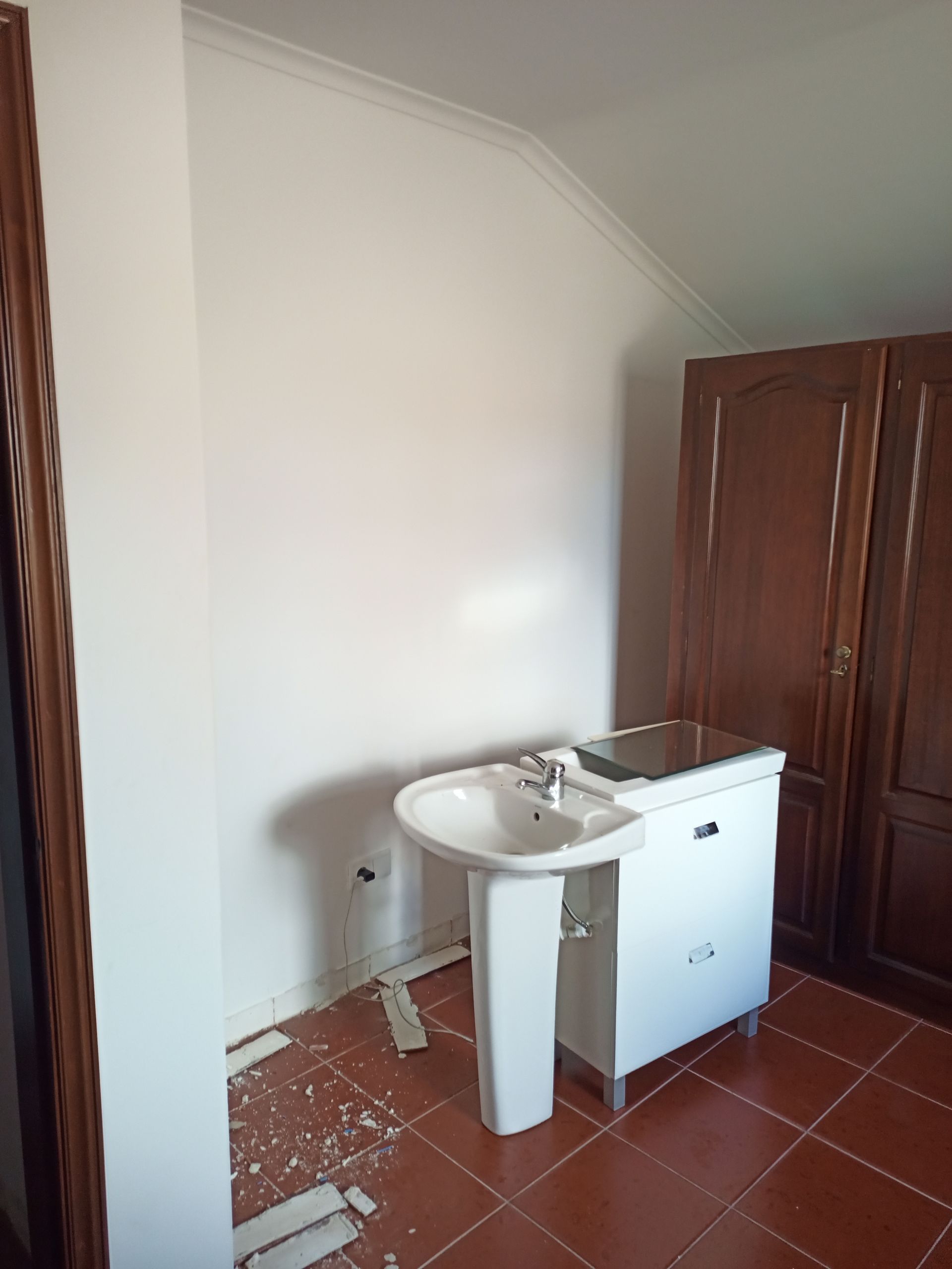 Banheiro com pia branca, armários e um guarda-roupa alto de madeira, sobre piso de azulejo.