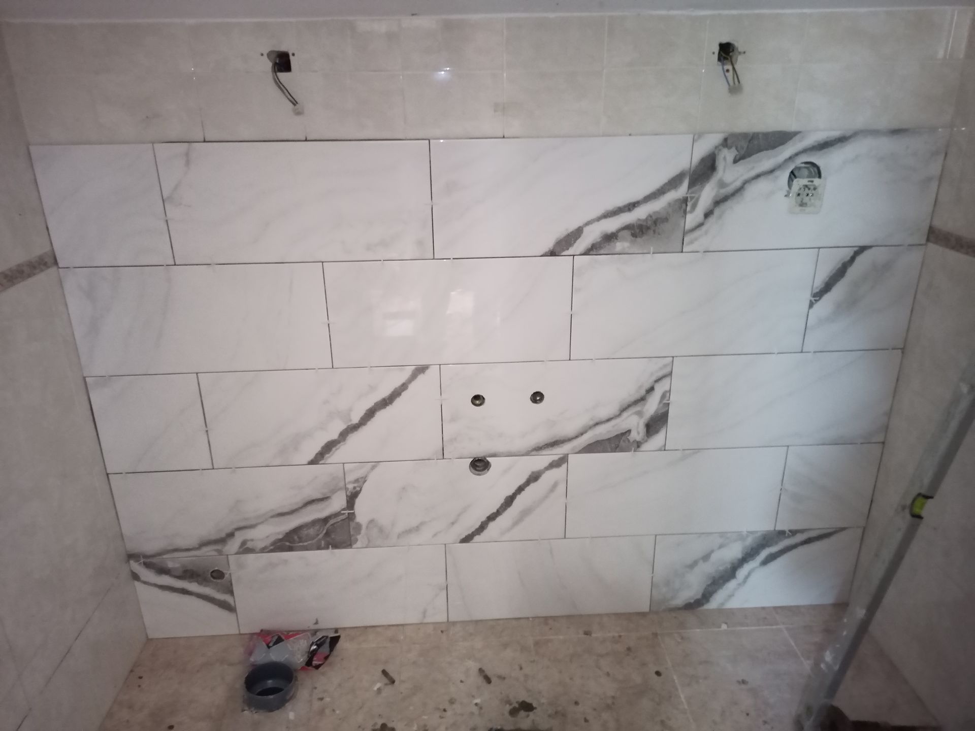 Azulejos brancos com estampa de mármore sendo instalados na parede de um banheiro, com tomadas elétricas visíveis.
