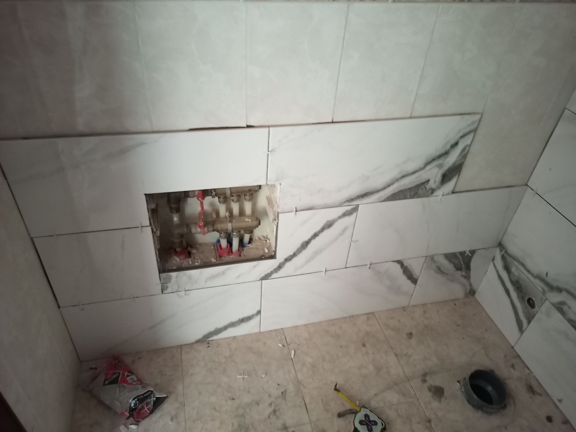 Azulejos parcialmente instalados na parede do banheiro, com um buraco expondo os canos; entulho de construção visível.