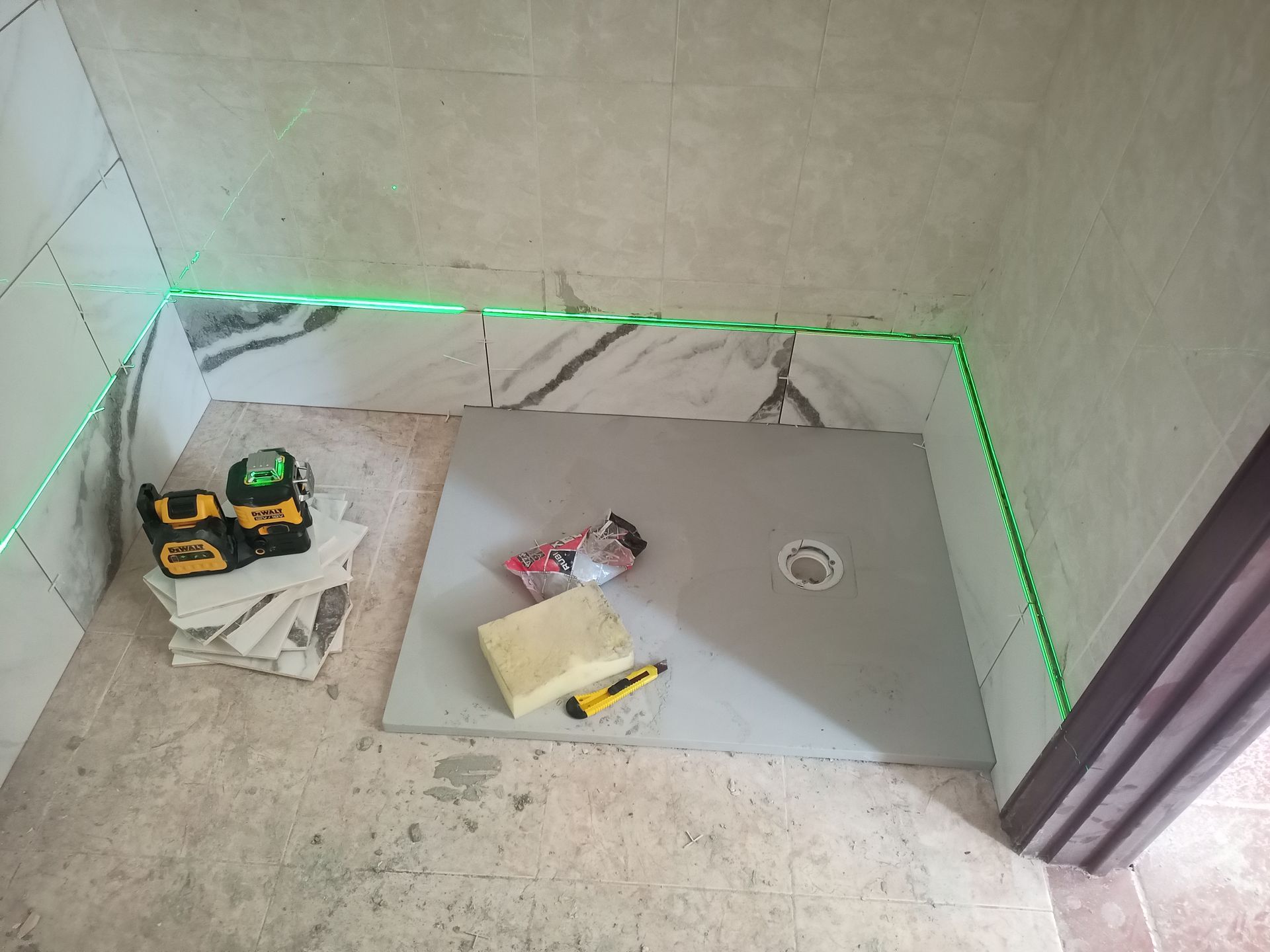 Linhas verdes de nível a laser marcando o layout dos azulejos em um banheiro. Base de chuveiro cinza instalada, ferramentas e sobras de azulejo por perto.