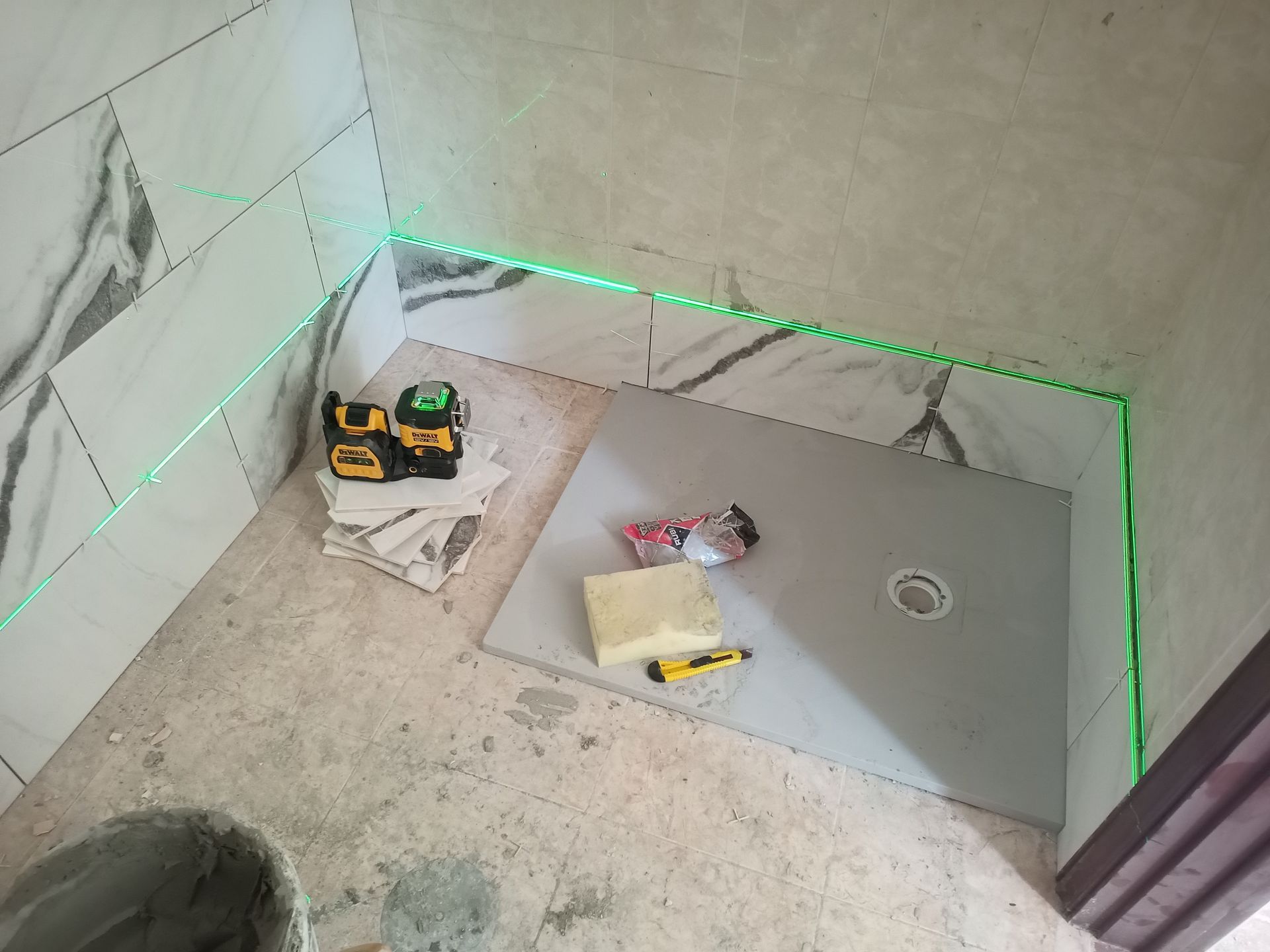 Box de chuveiro em construção, com azulejos imitando mármore, base de chuveiro cinza e um nível a laser emitindo linhas verdes.
