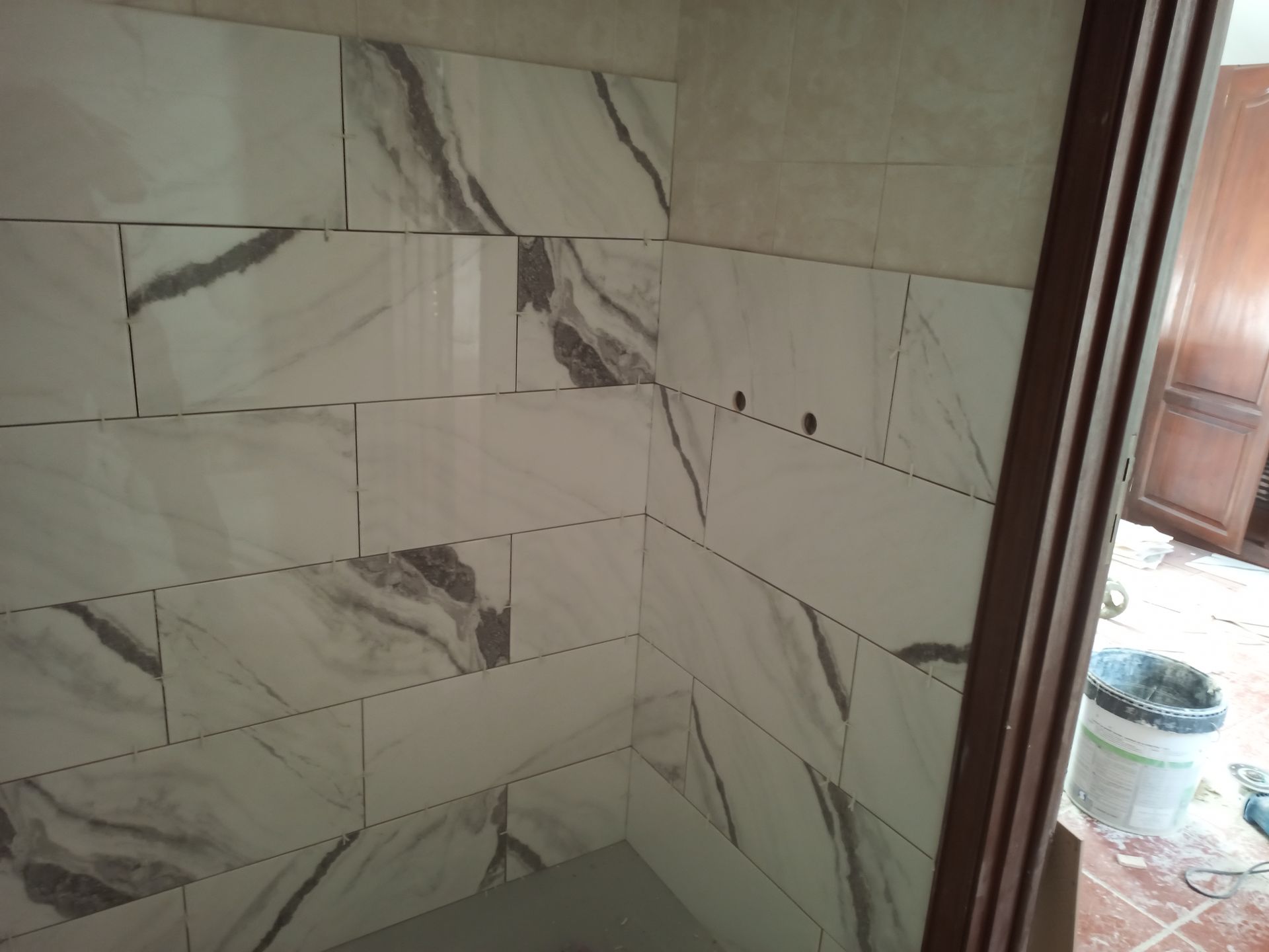 Paredes do banheiro revestidas com azulejos que imitam mármore branco e cinza. Batente da porta marrom visível.