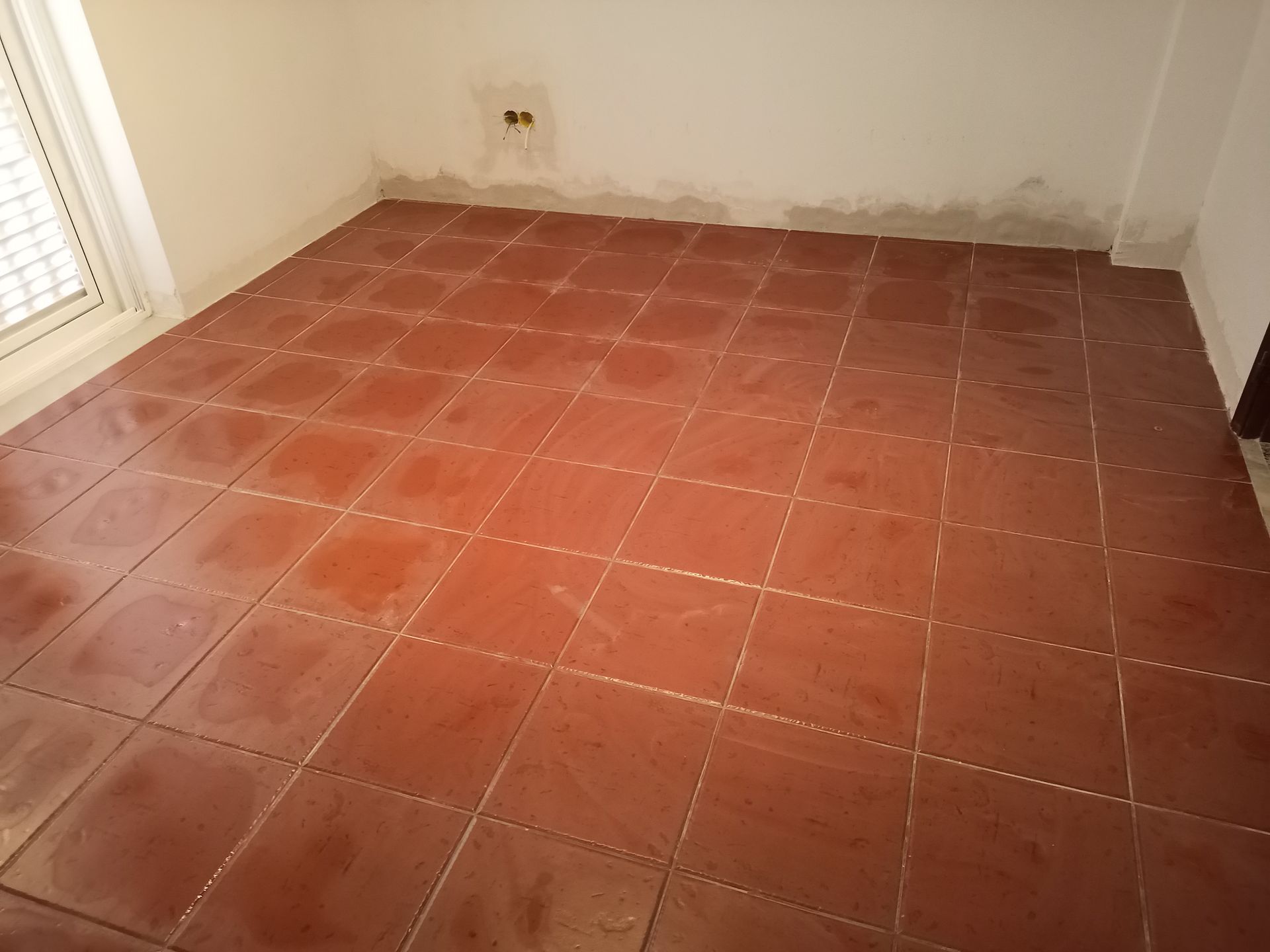 Piso de azulejos marrons em um cômodo com paredes brancas, próximo a uma janela.