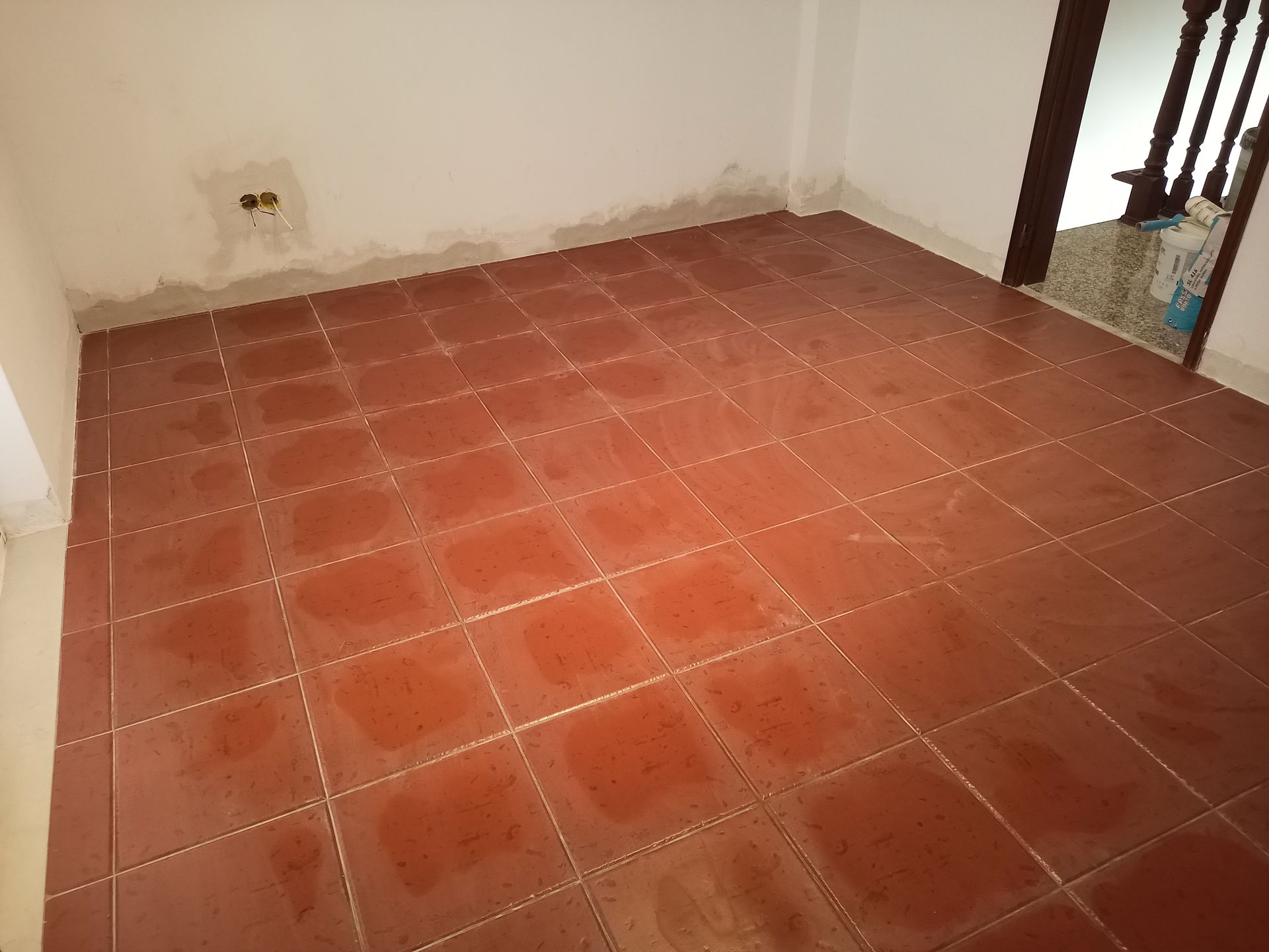Quarto com piso de azulejo vermelho e paredes brancas. Batente da porta marrom visível. Tomada elétrica na parede.