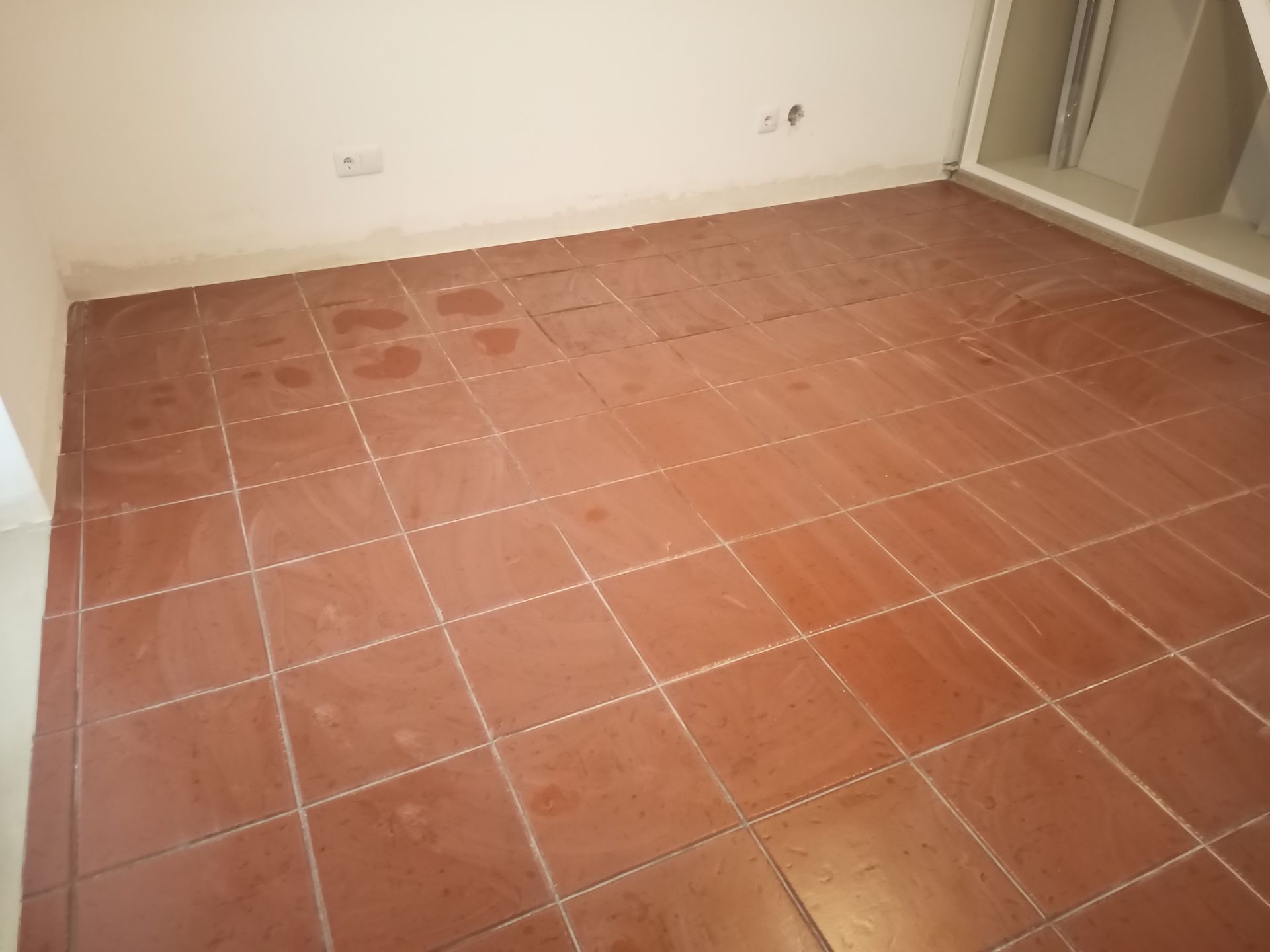 Piso de ladrilho de terracota em um cômodo com paredes brancas e uma abertura de janela parcialmente visível.