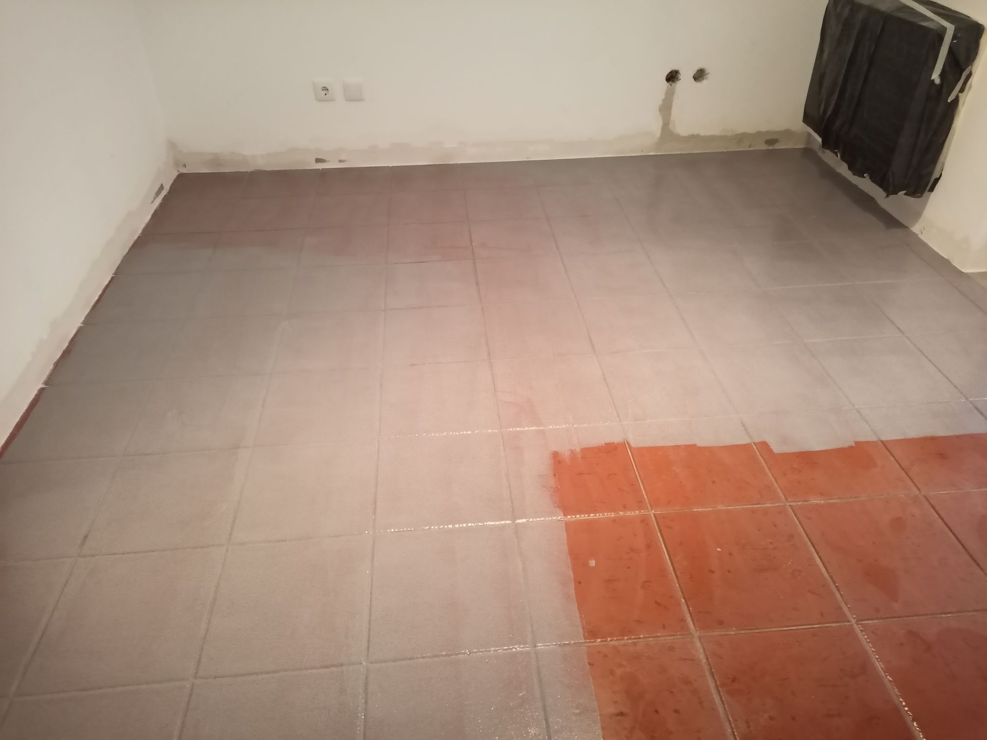 Quarto com piso de azulejo parcialmente pintado de cinza, com alguns detalhes em vermelho visíveis. Paredes brancas, aquecedor à direita.